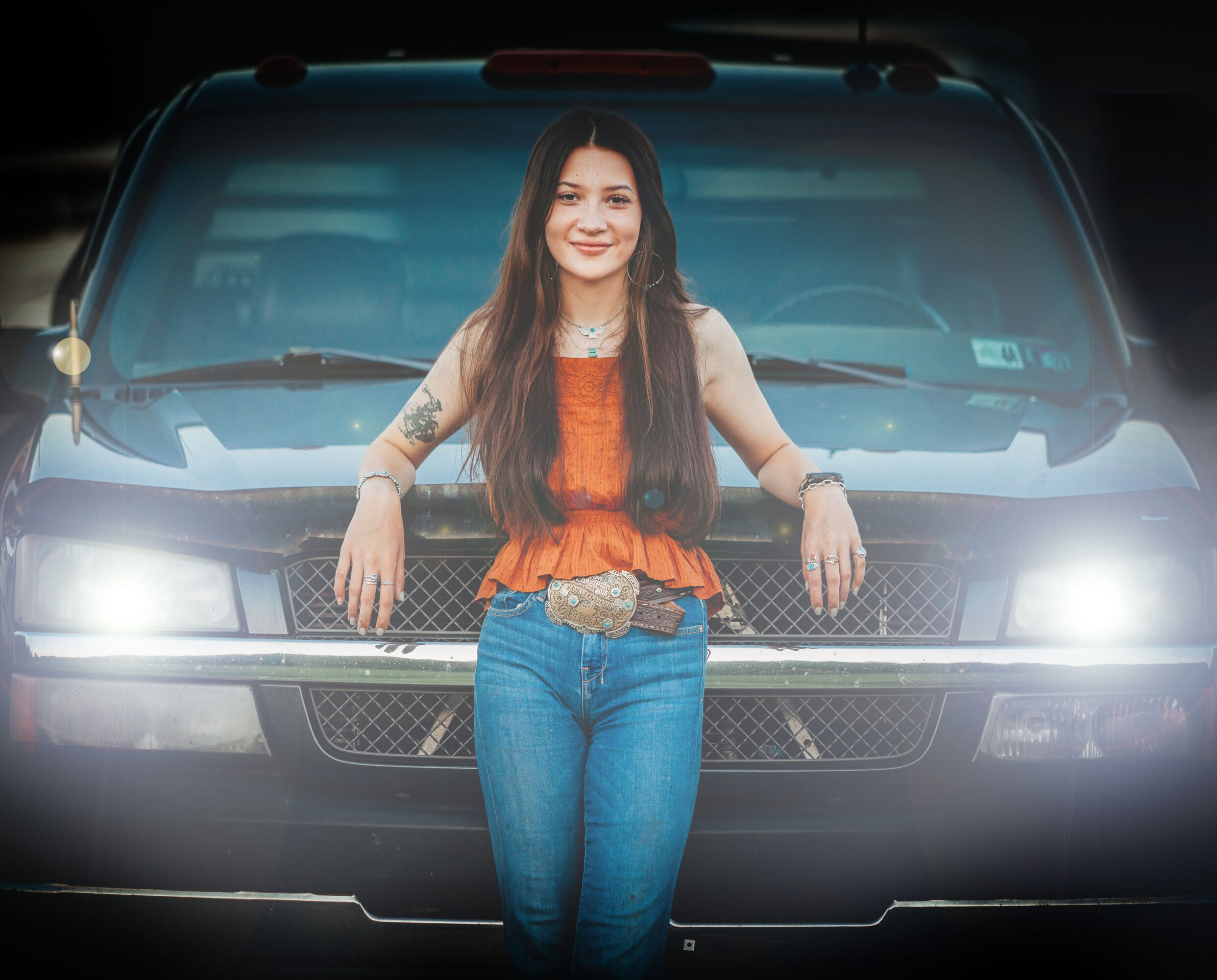 Young woman with long hair standing in front of a vehicle, smiling, wearing a sleeveless orange top and jeans.