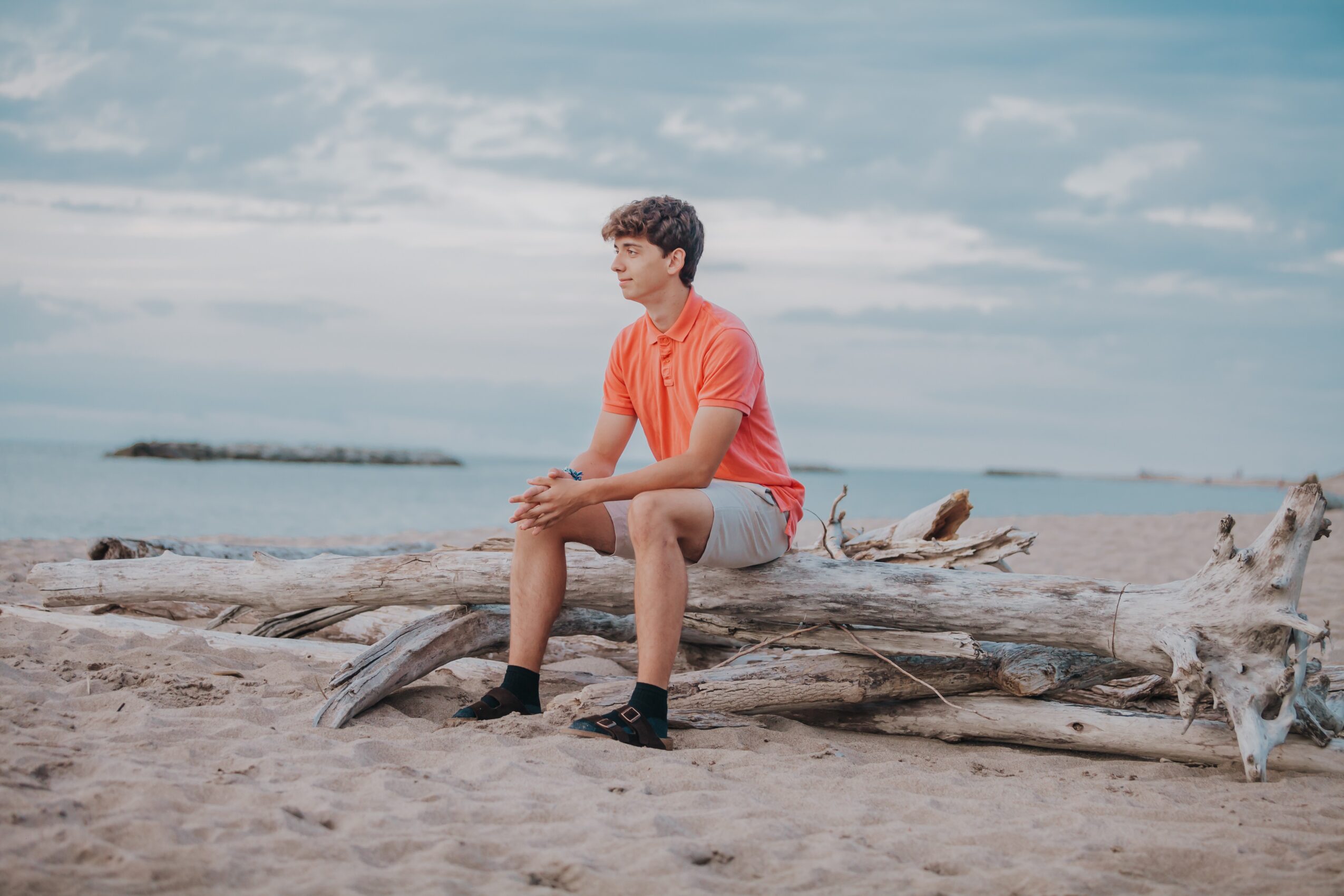 Young person sitting on driftwood at the beach, looking into the distance, with a cloudy sky and ocean in background.