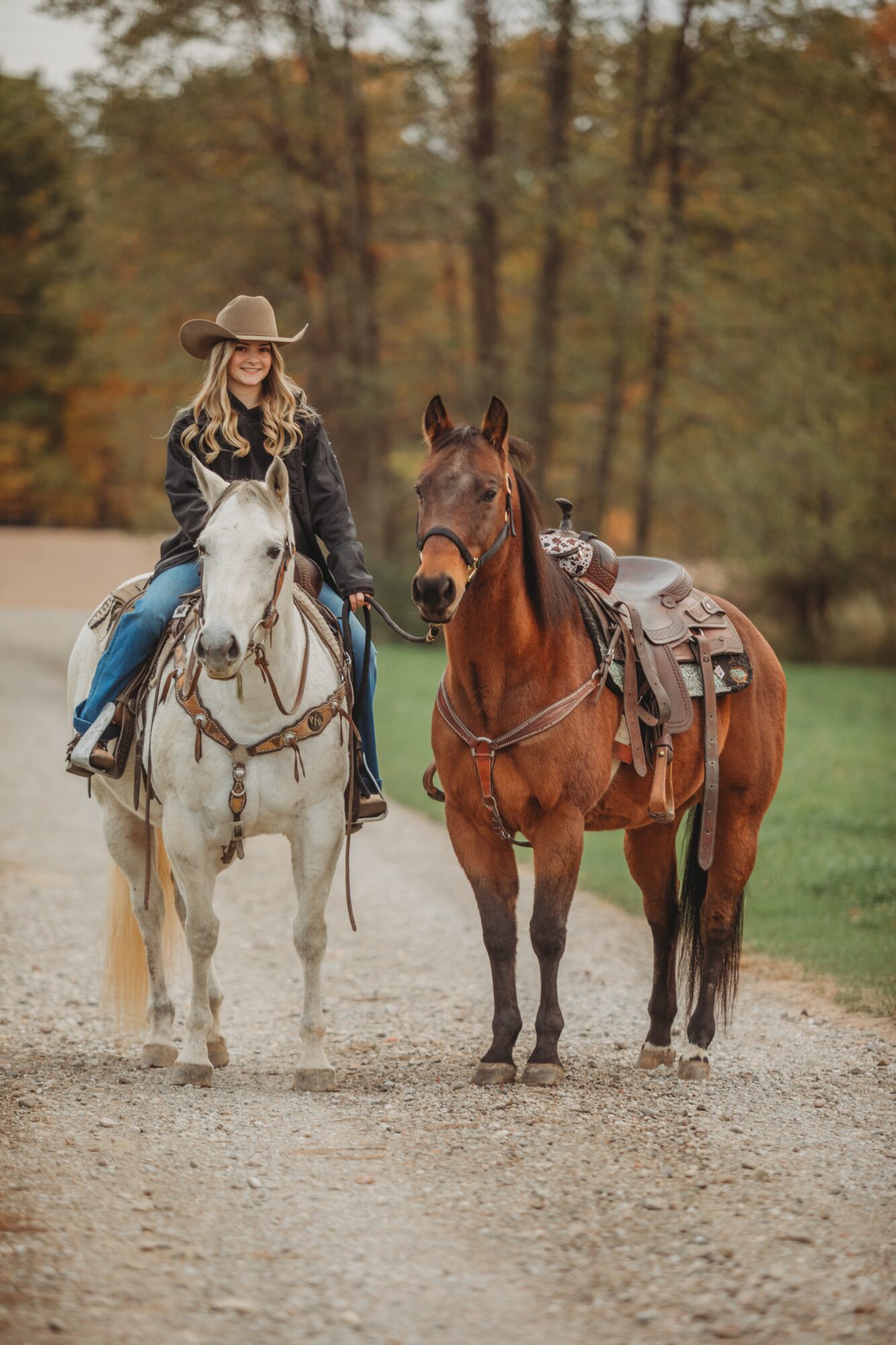 Woman in cowboy hat riding a white horse, standing next to a brown horse with saddle, on a dirt path in autumn forest.