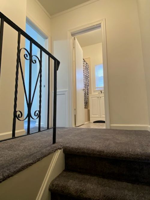 View of a staircase with a black metal railing, leading to a doorway opening into a room with a window and patterned curtain.