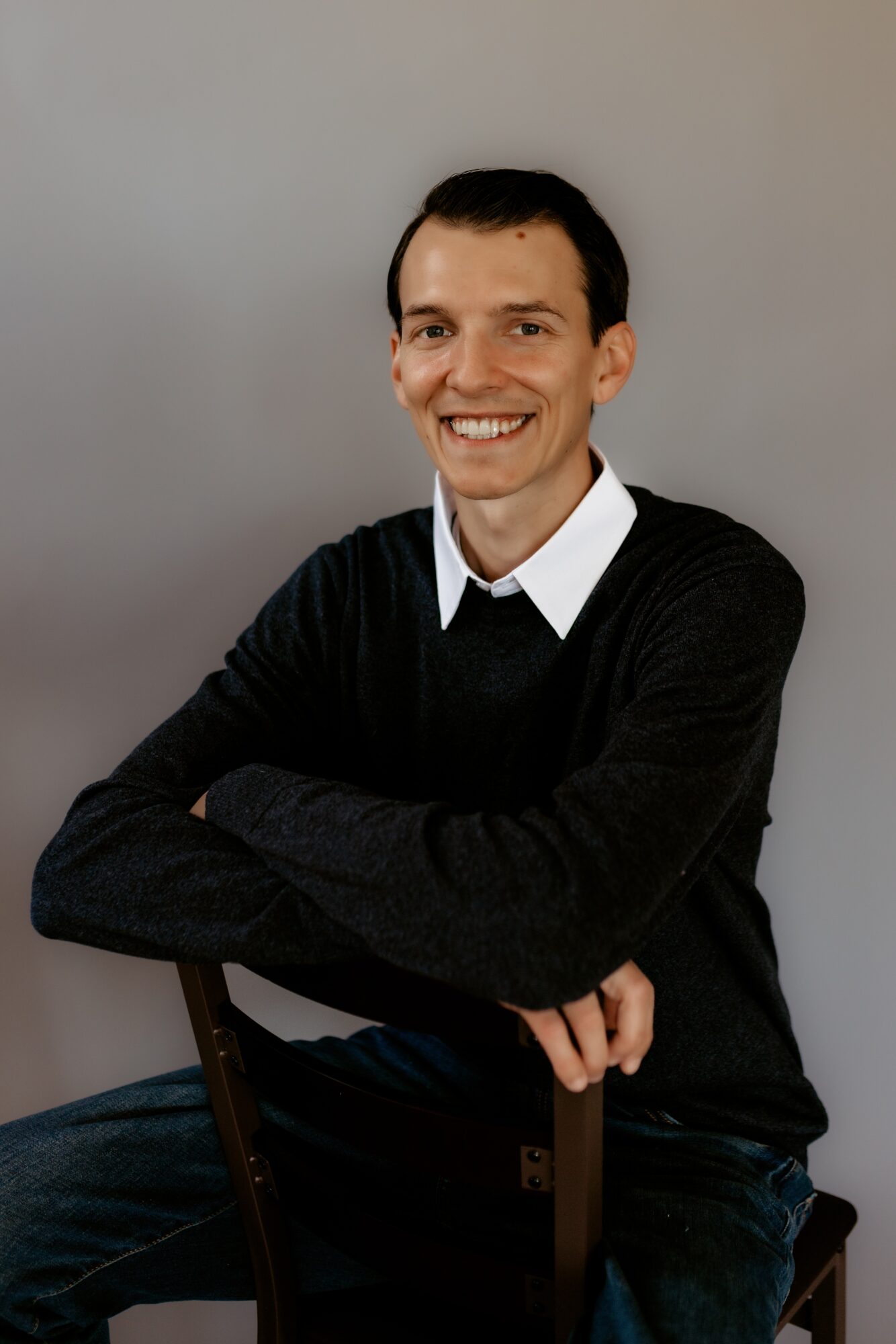 Smiling man with dark hair, wearing a black sweater over a white shirt, sitting with arms crossed against a plain background.