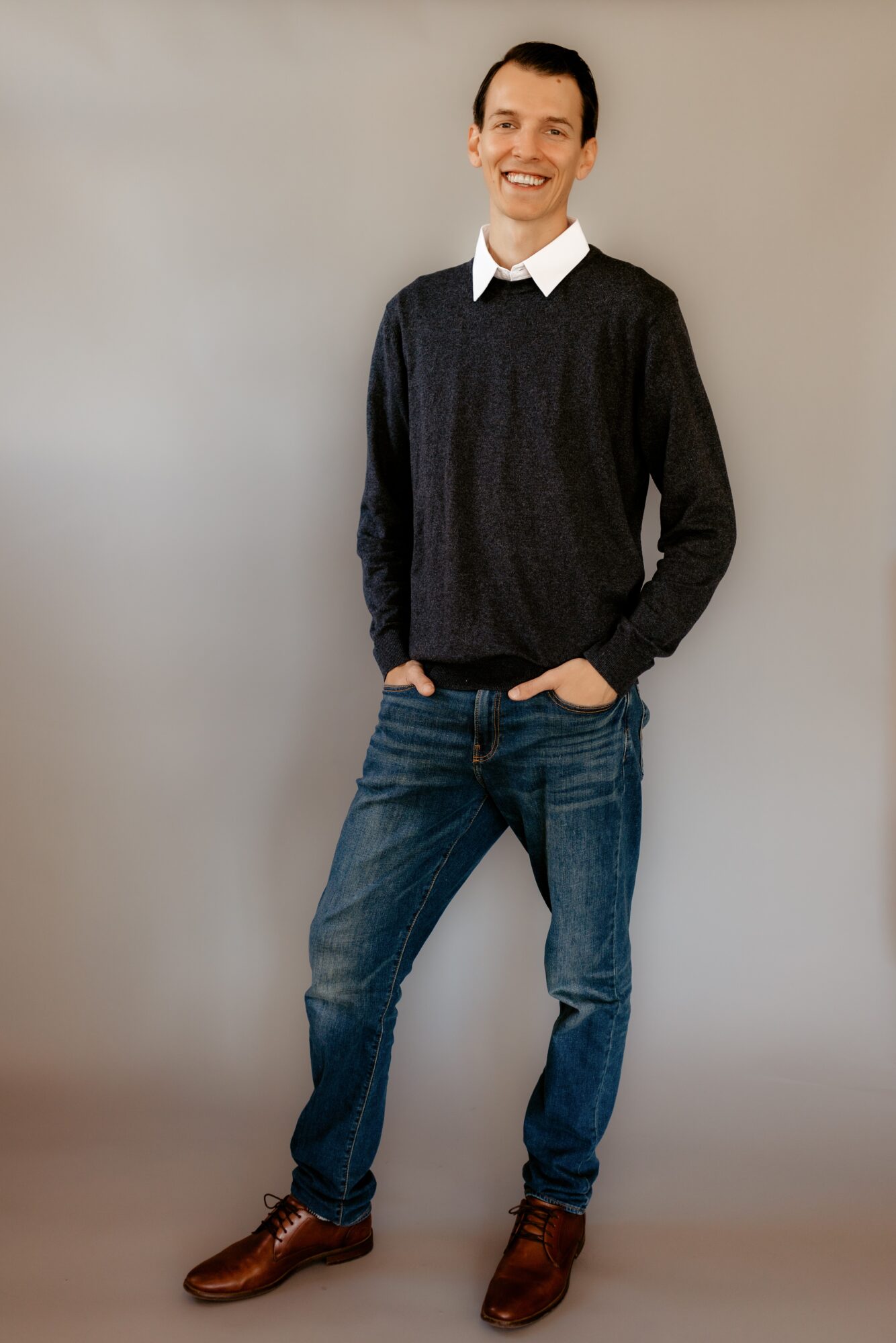 Young man standing with hands in pockets, smiling, wearing a dark sweater, white shirt, jeans, and brown shoes.