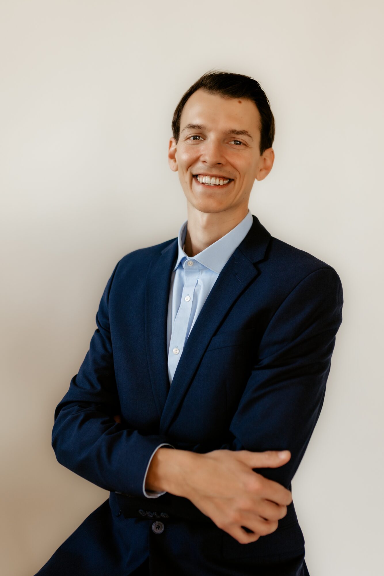 Man in a dark suit and light blue shirt smiling with arms crossed against a plain background.
