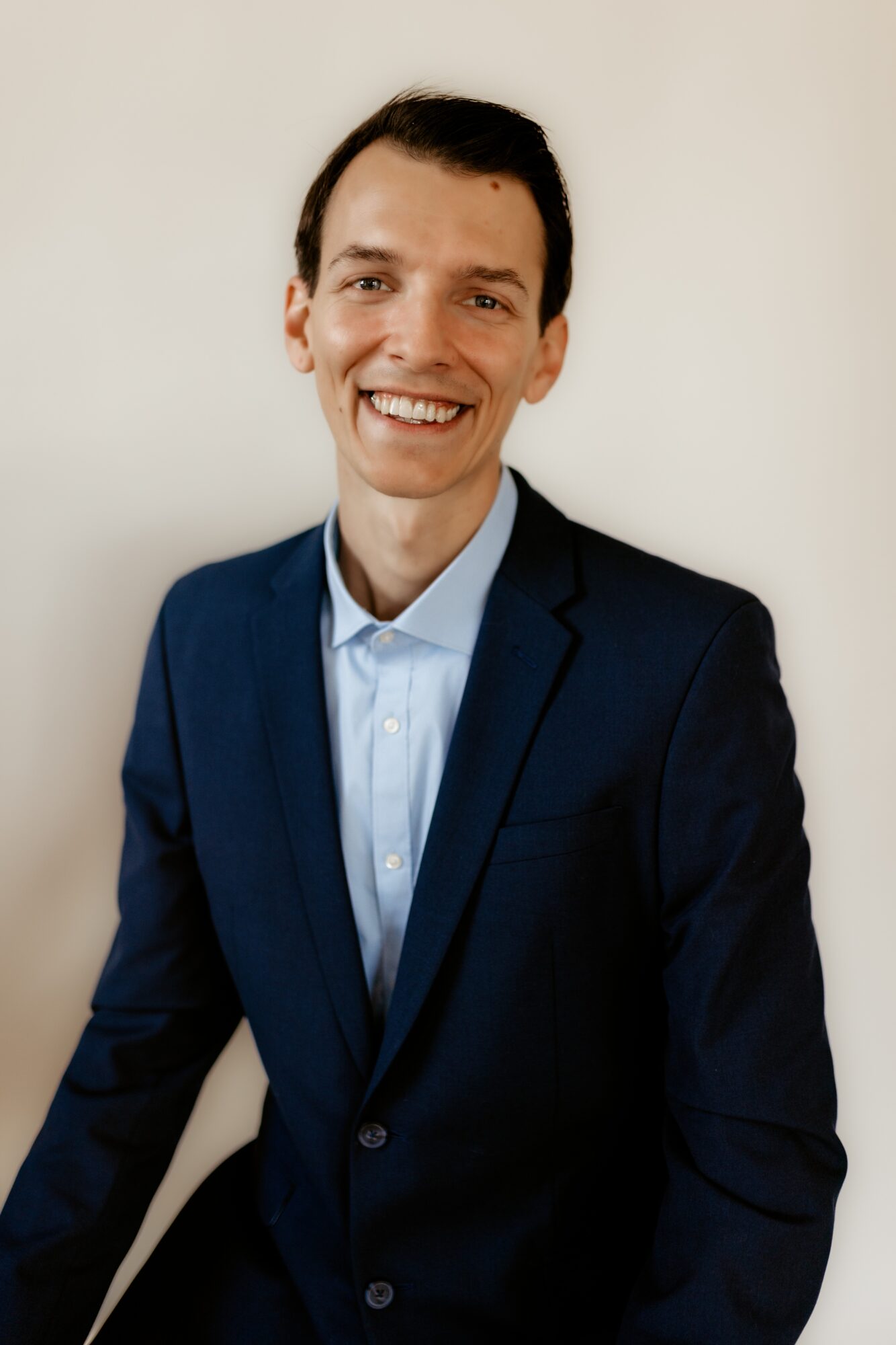 Man in a dark suit and light blue shirt smiling, standing against a plain background.