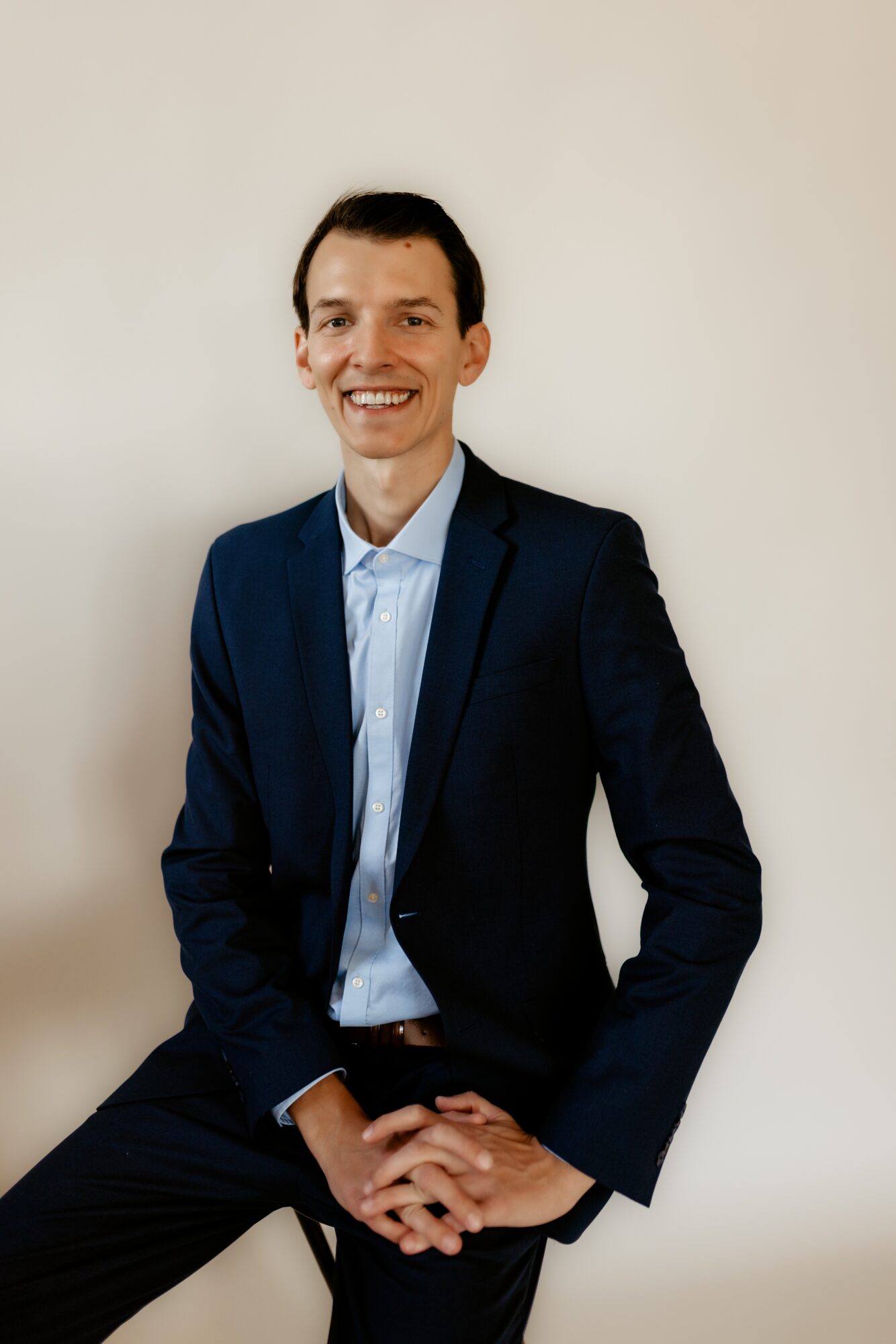 Man in a dark suit and light blue shirt sitting against a plain light background, smiling.