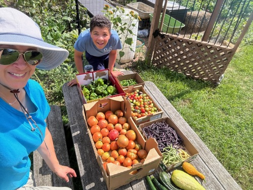 Two people smiling outdoors with a wooden table of fresh fruits and vegetables, garden and fence in background.