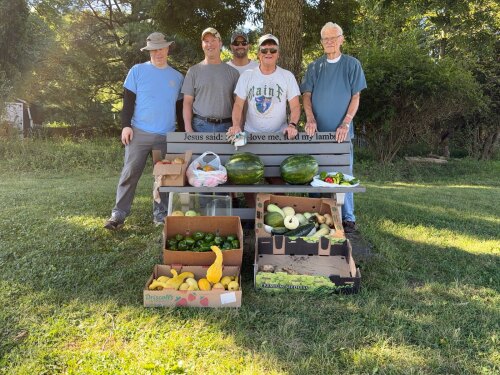 Four people stand behind boxes of watermelons, cucumbers, and yellow squash on grass in a park with trees in the background.