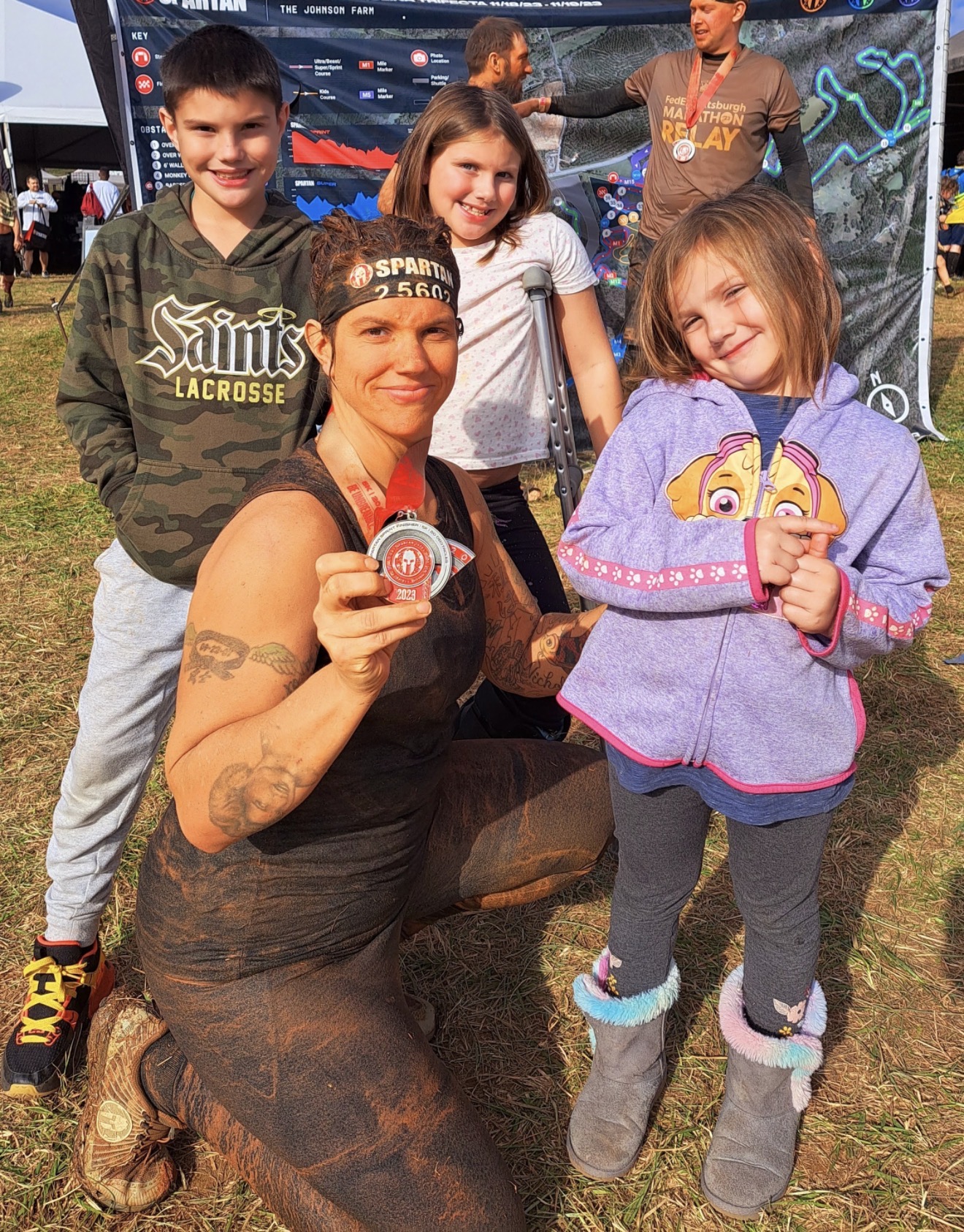 Group of children and a woman outdoors, smiling, with a large screen in the background, some holding medals.