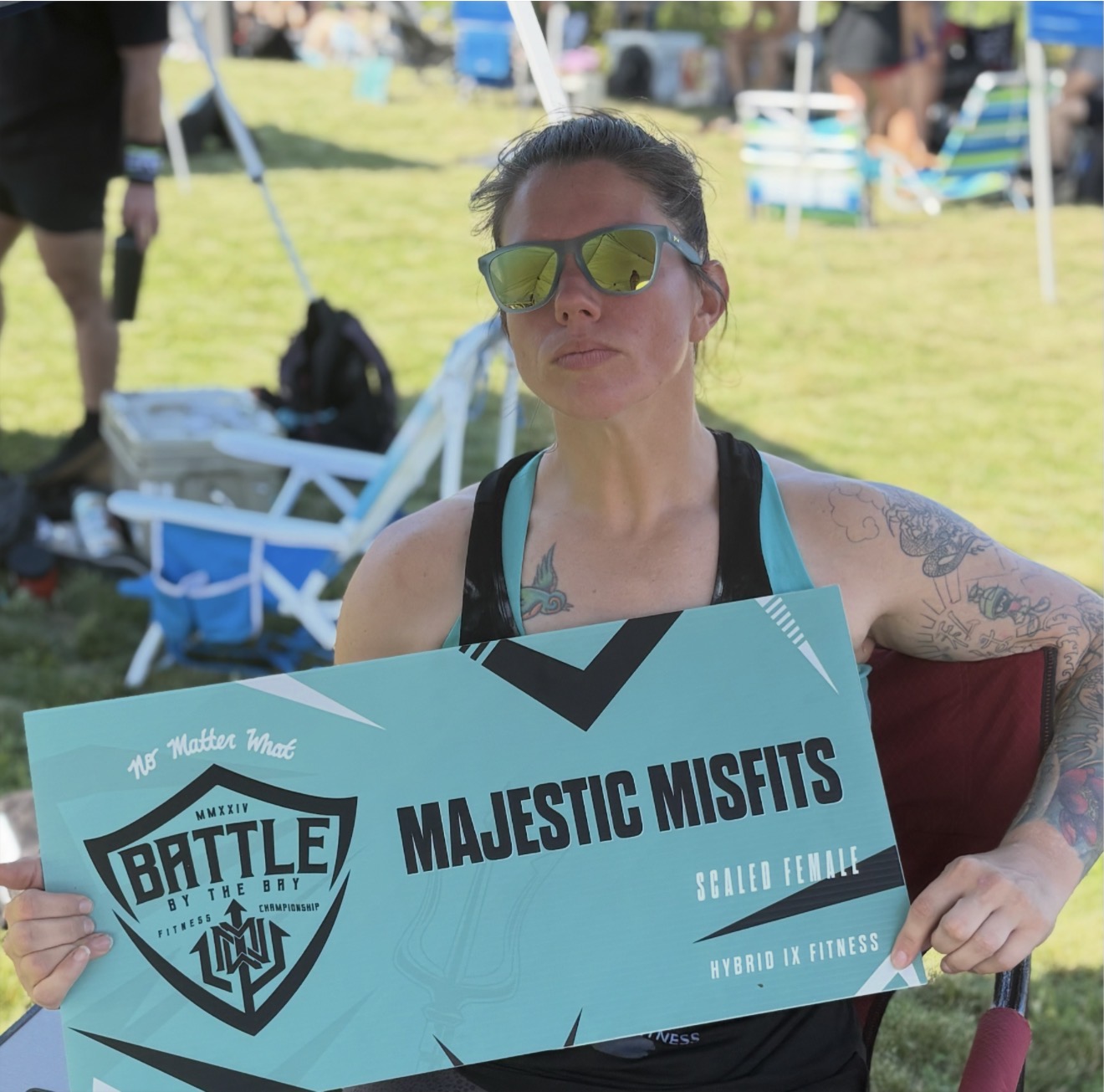 Woman holding a sign at an outdoor event, wearing sunglasses and a tank top, with tents and chairs in the background.