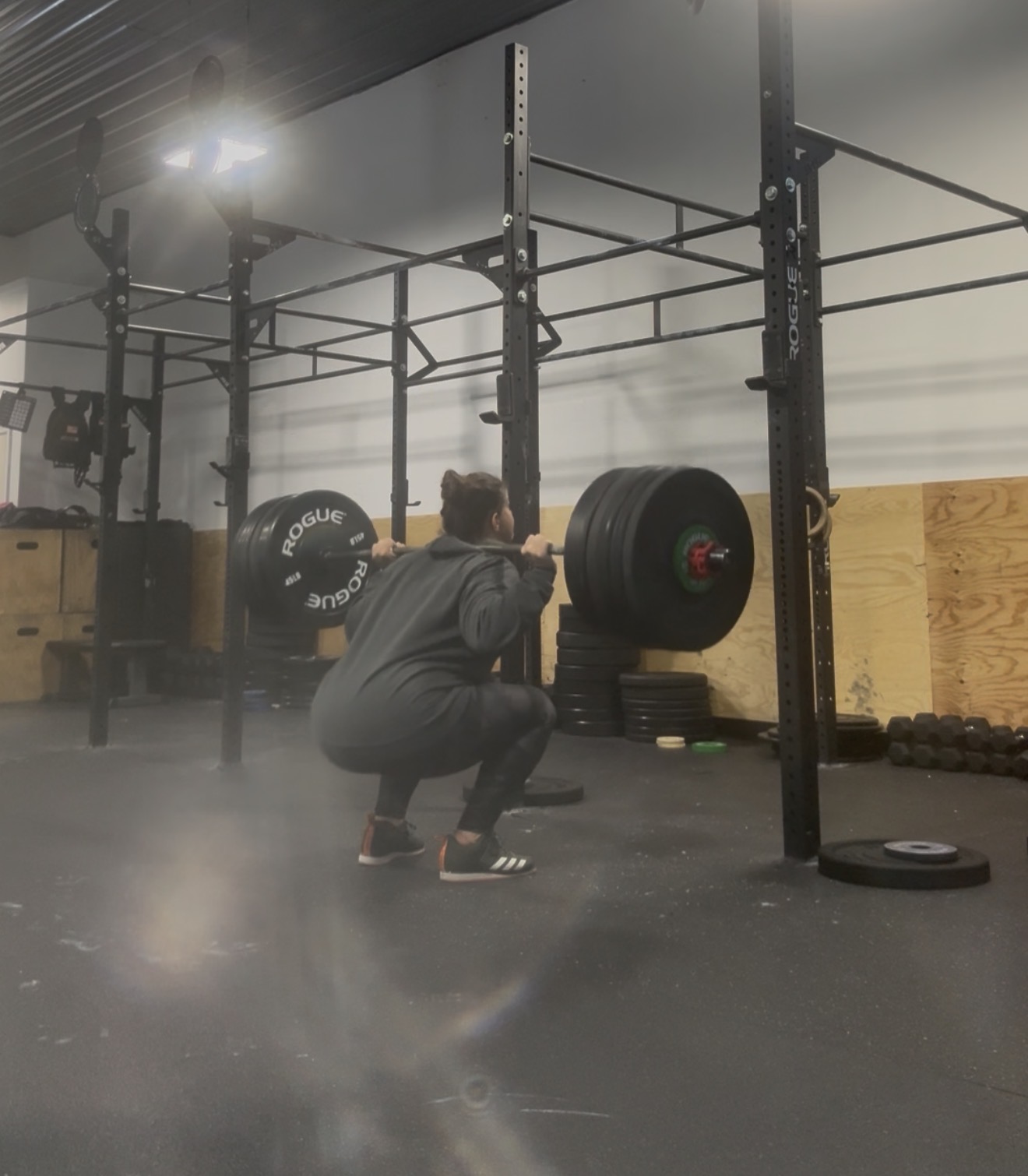 Person squatting in a gym near a barbell with large weights, black flooring, and gym equipment in the background.