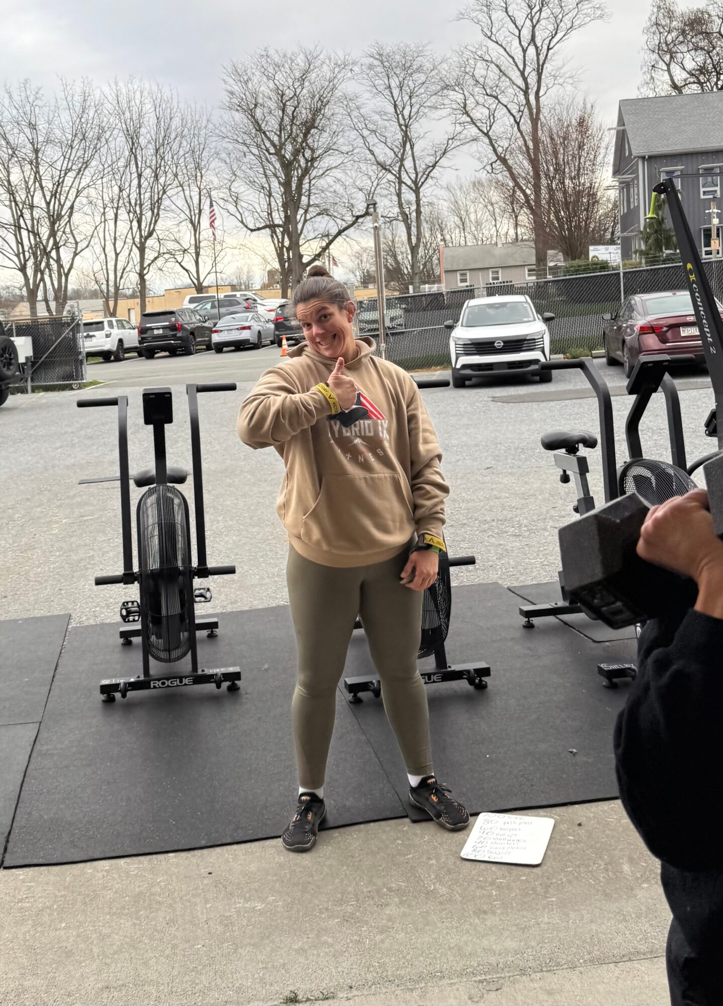 Person drinking from a cup in a gym parking lot with exercise equipment and parked cars behind.