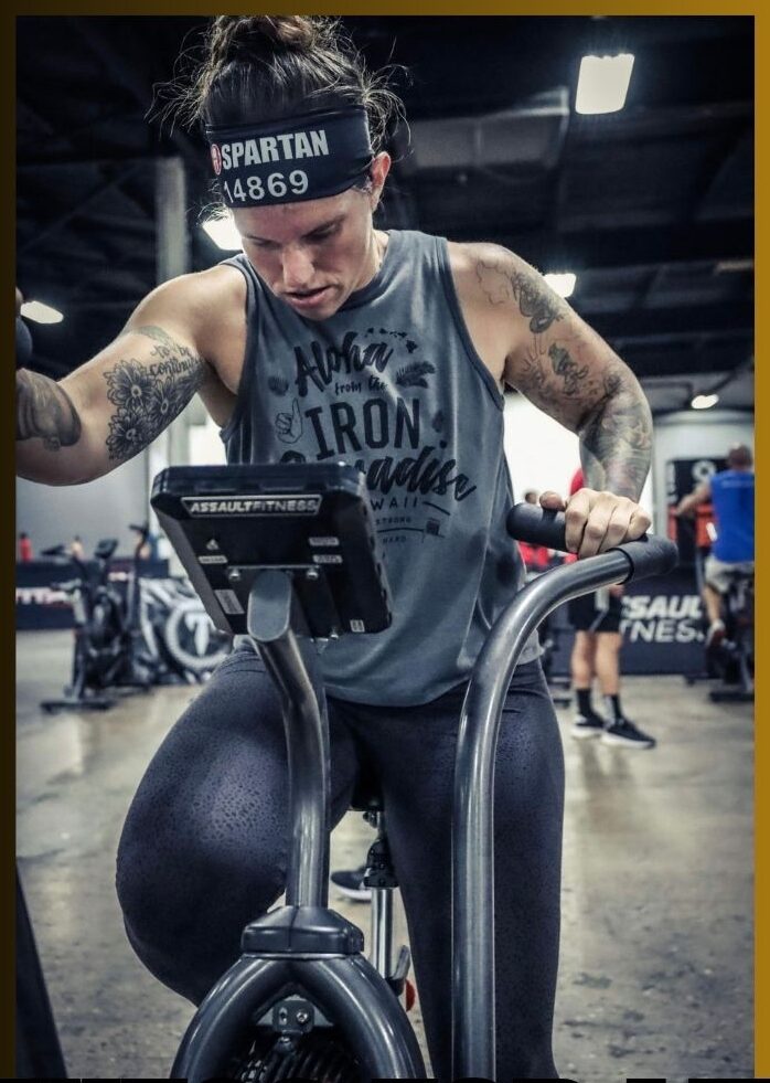 Woman with tattoos on arms, wearing a headband and workout shirt, using exercise bike in gym, looking at screen.