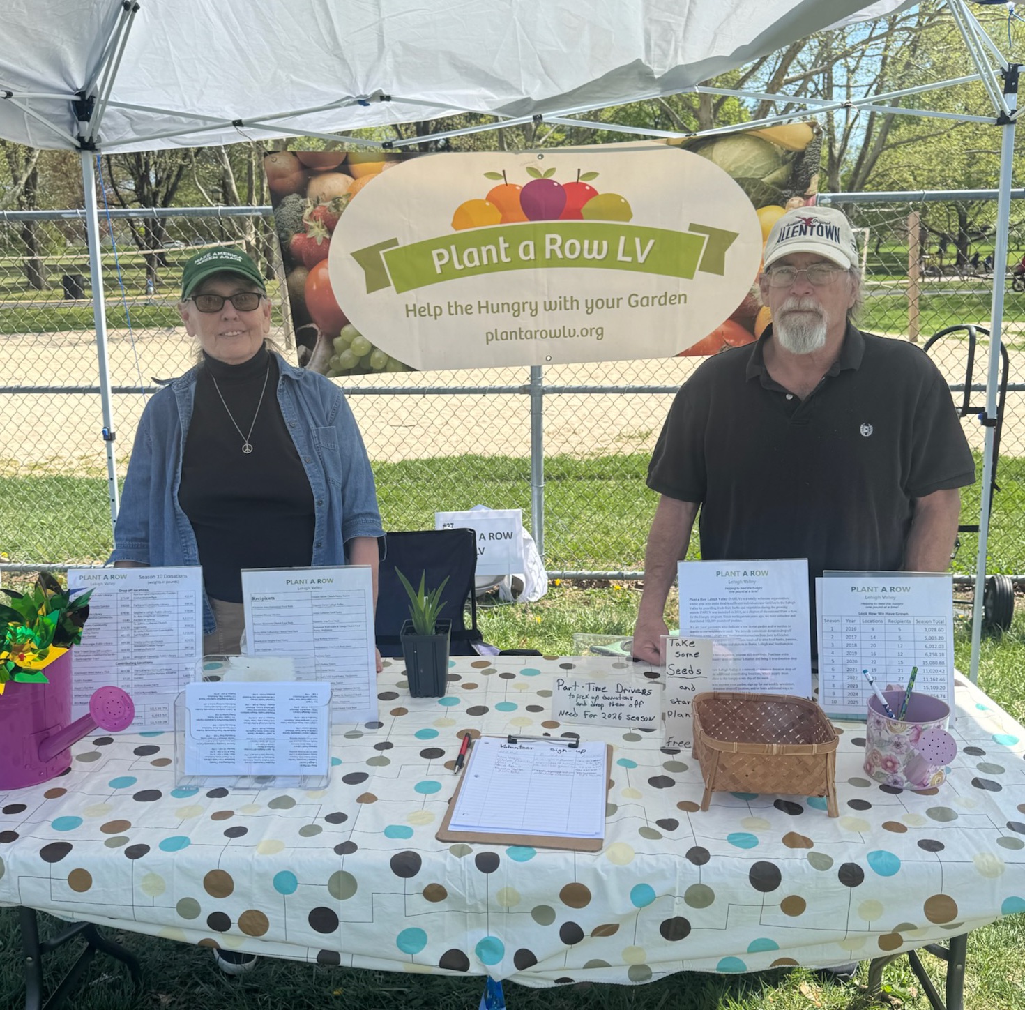 Two people stand behind a table at a garden event, with a sign reading 'Plant a Row V' and various papers on the table.