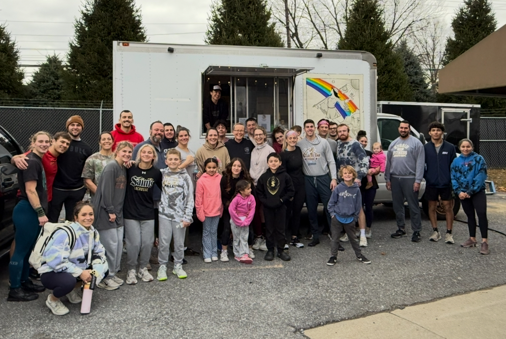 Group of people standing in front of a food truck outdoors, some children and adults smiling, trees in background.