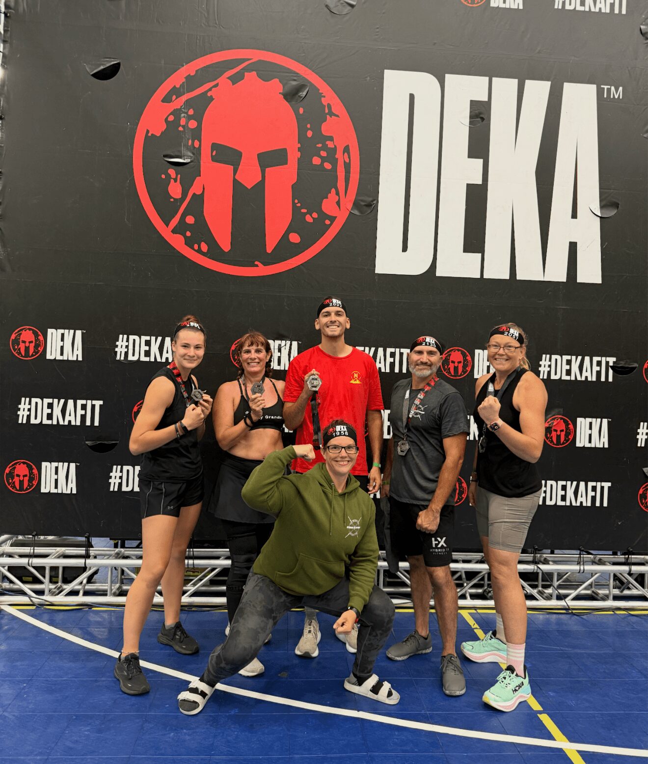 Group of six people posing in front of a large DEKA logo and Spartan helmet symbol, on a blue sports court.