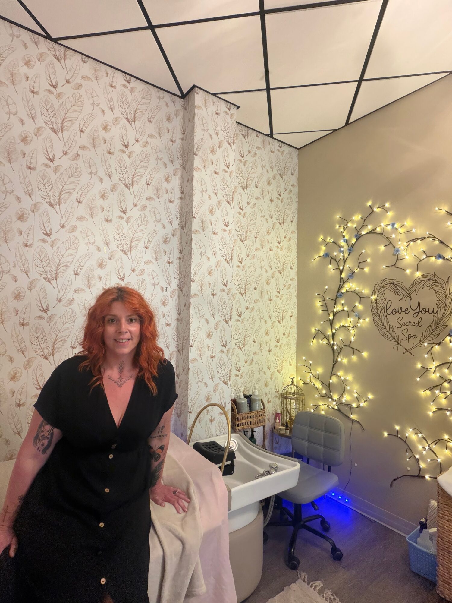 Woman with curly red hair standing indoors near a decorated wall with string lights and a tree design, smiling.