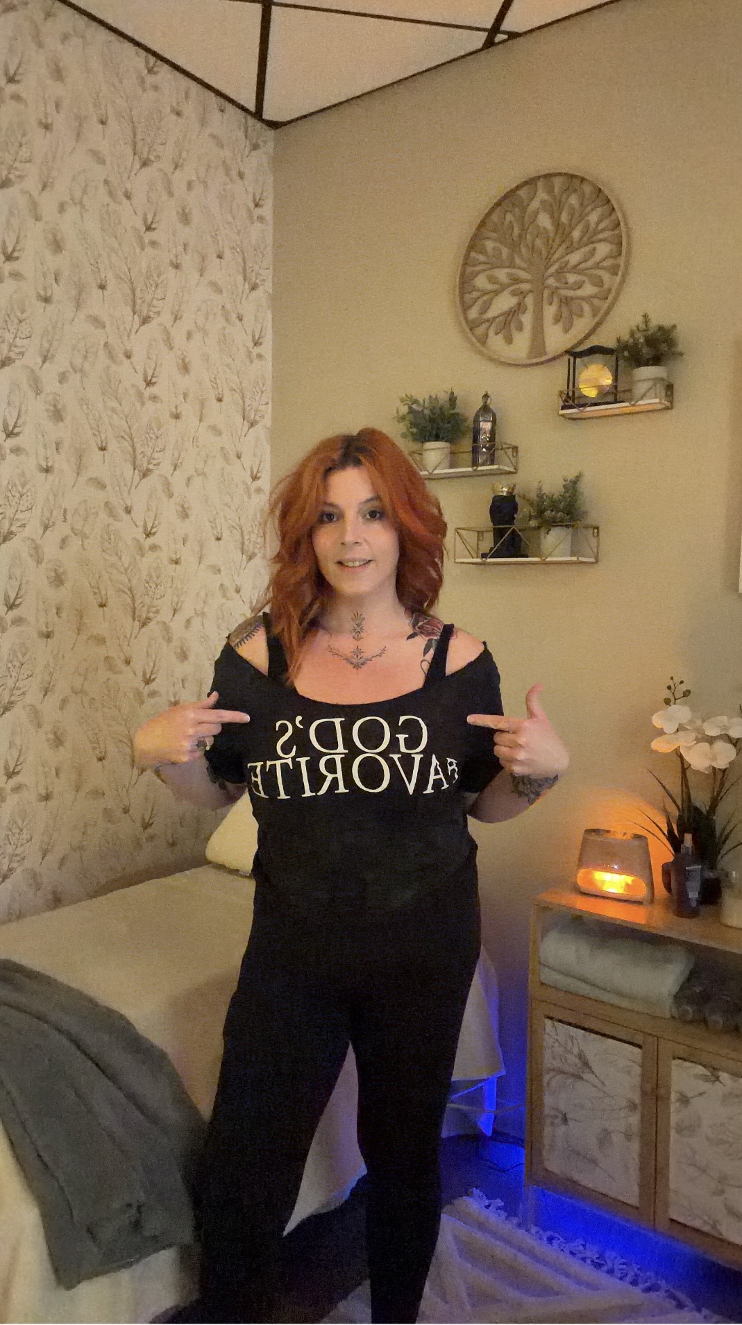 Woman with red hair standing indoors, pointing at a sign, with decorative shelves and plants behind her.