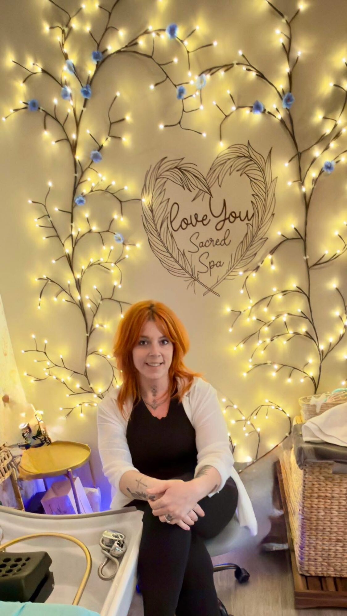 Woman with red hair sitting in front of illuminated decorative branches and a heart-shaped sign with handwritten message.