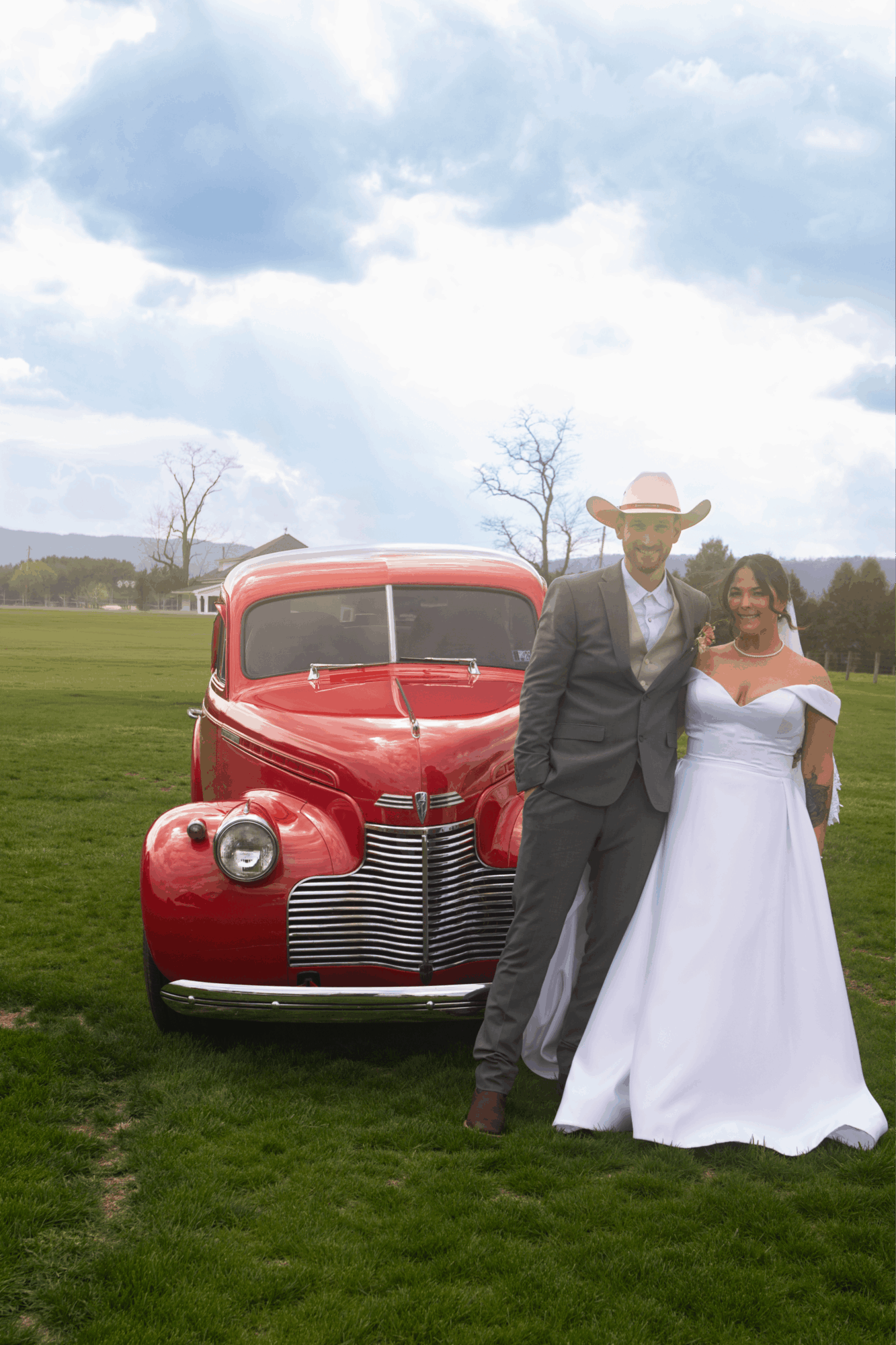 A man in a suit and cowboy hat stands next to a woman in a white dress, with a red vintage car behind them outdoors.