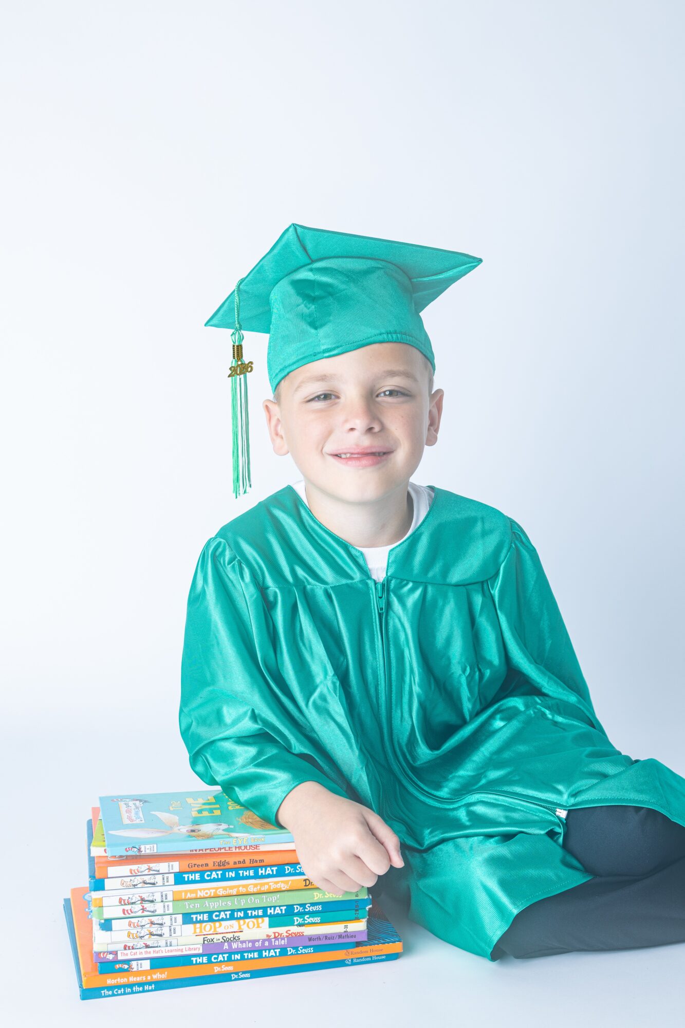 Young child in graduation cap and gown sitting next to a stack of books, smiling, against a plain background.