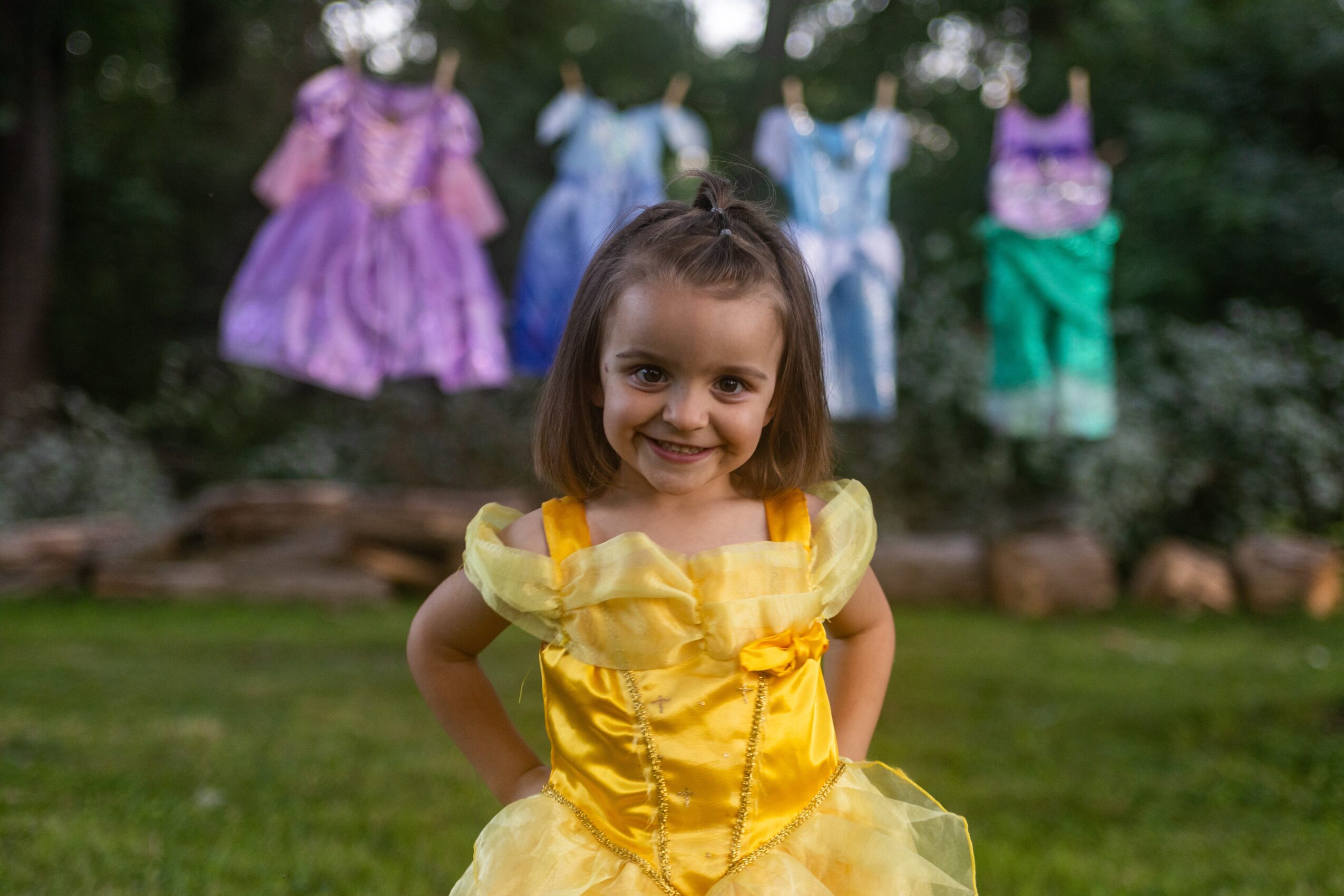 Smiling girl in a yellow dress stands outdoors with colorful dresses hanging in the background.