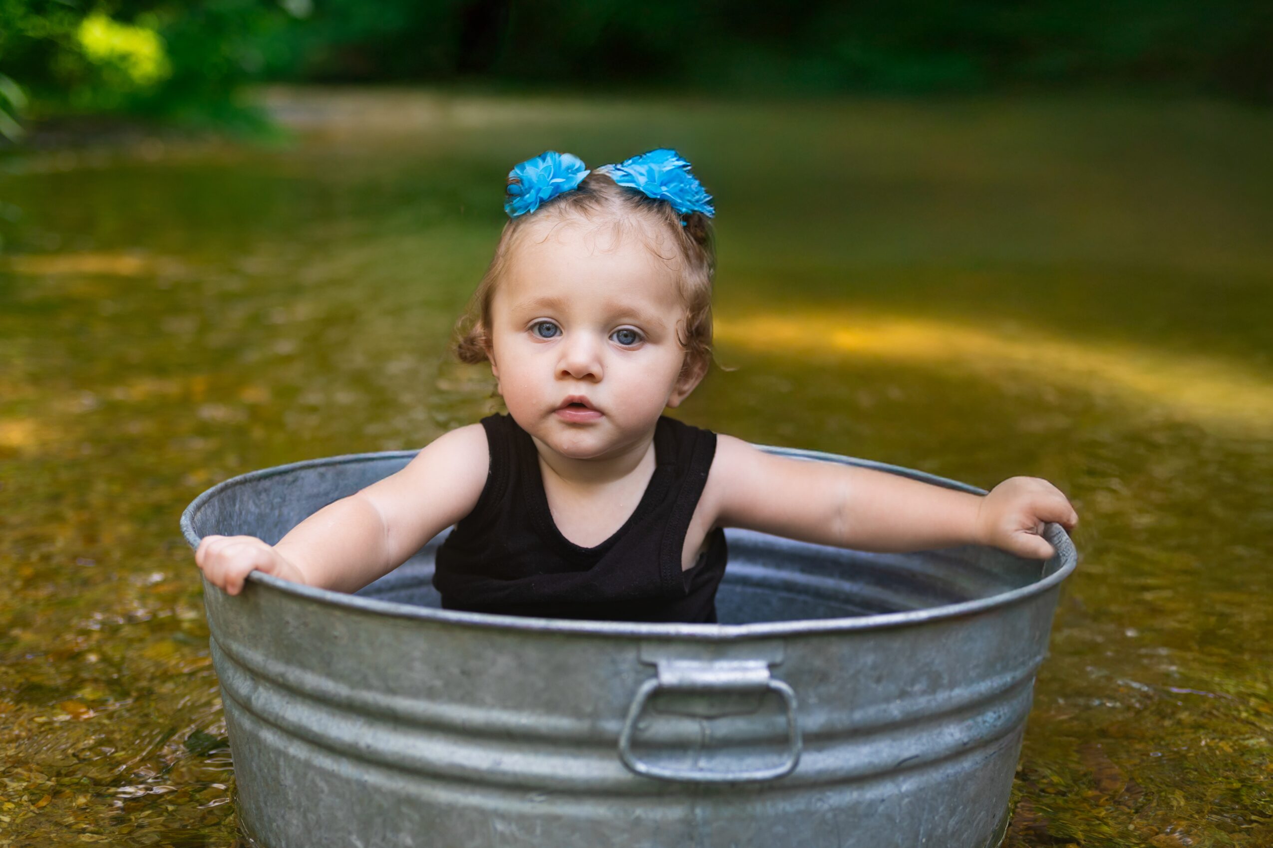 Child with blue bows in hair sitting in a metal tub in water outdoors, with trees in background.