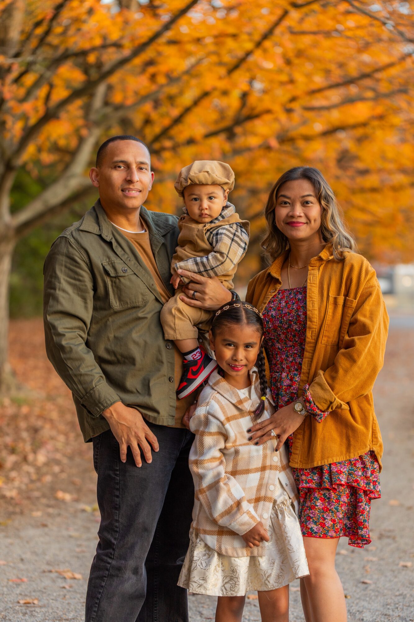 Family of four outdoors in autumn, trees with orange leaves in background, smiling, standing on a path.