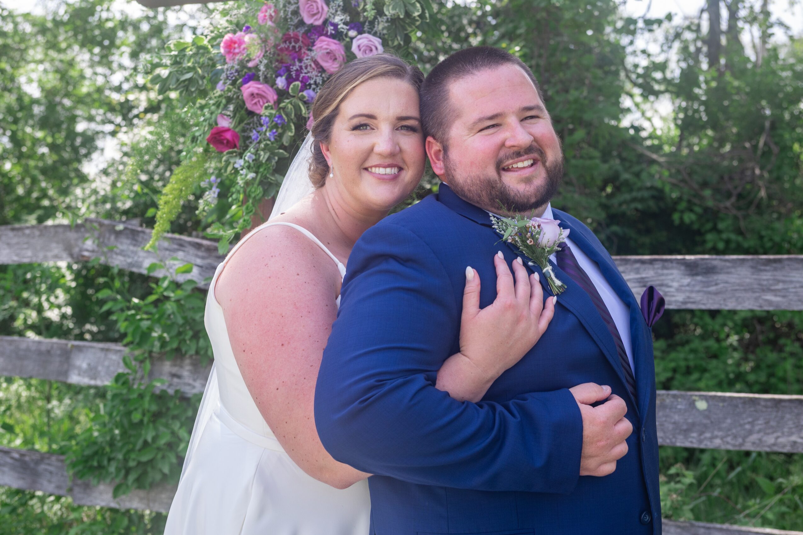 Smiling bride and groom embrace outdoors with greenery and a wooden fence in background.