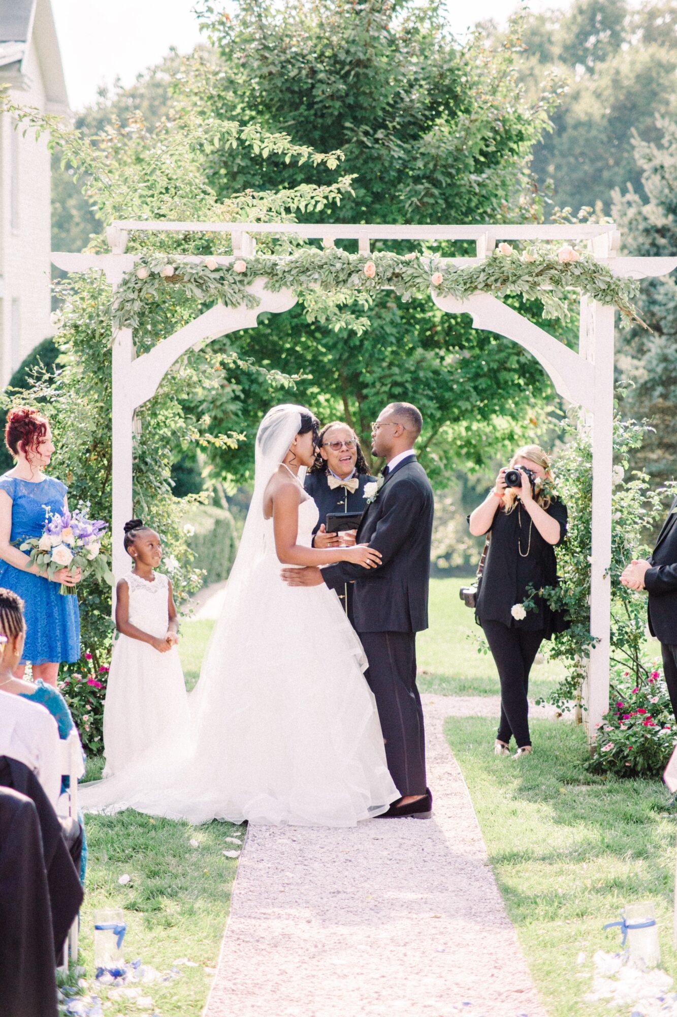 Bride and groom holding hands and smiling at each other under an arch during outdoor wedding ceremony, surrounded by guests and greenery.