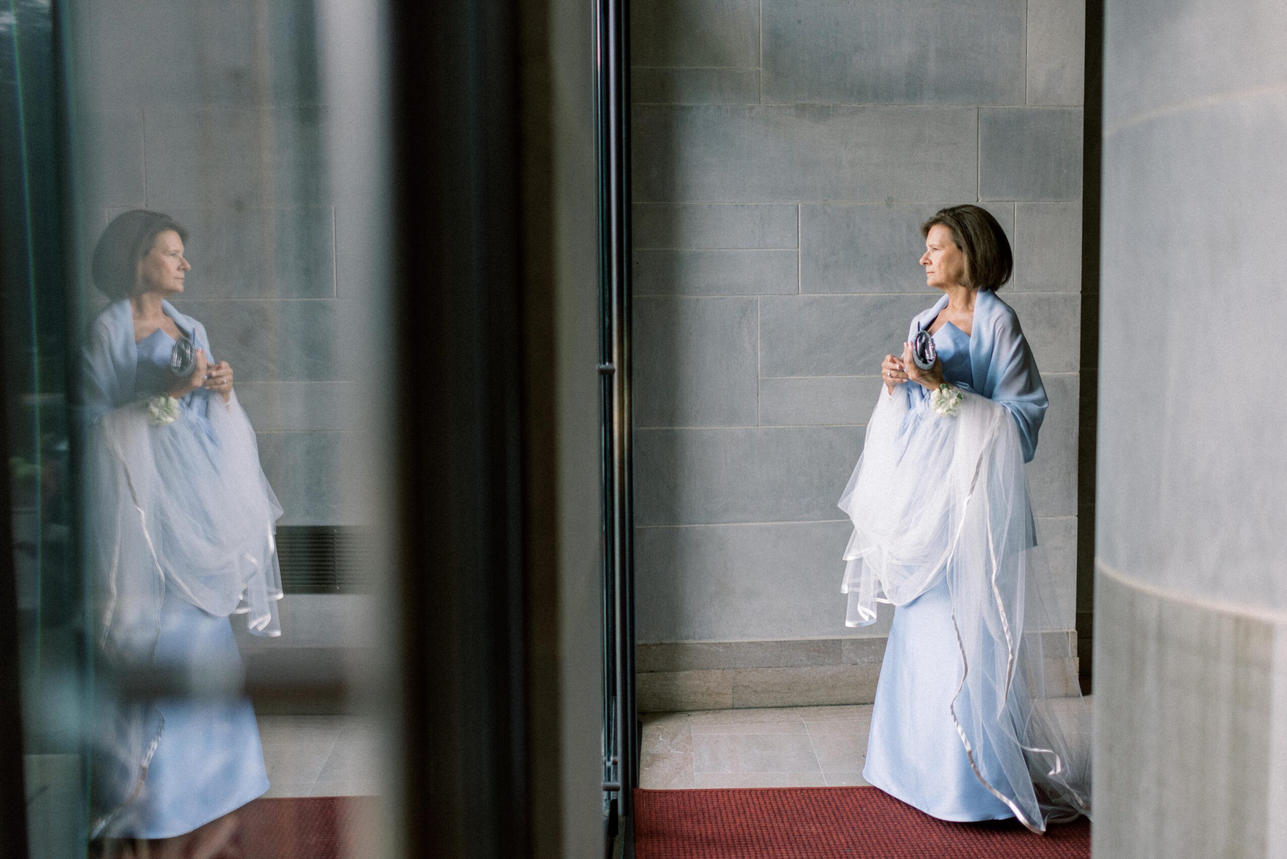 Woman in a light blue dress and white shawl standing against a gray wall, reflected in glass, holding a drink.