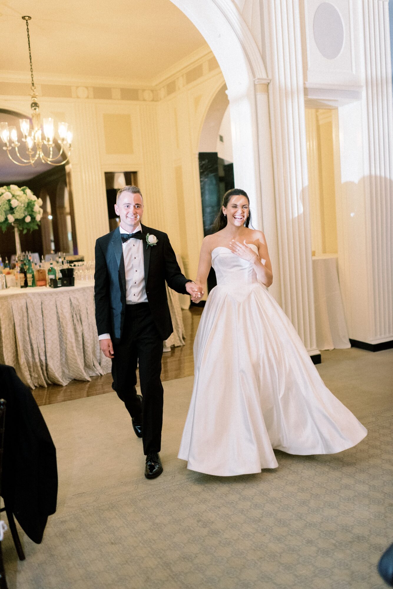 A bride and groom walk hand in hand in a decorated room with chandeliers and floral arrangements.