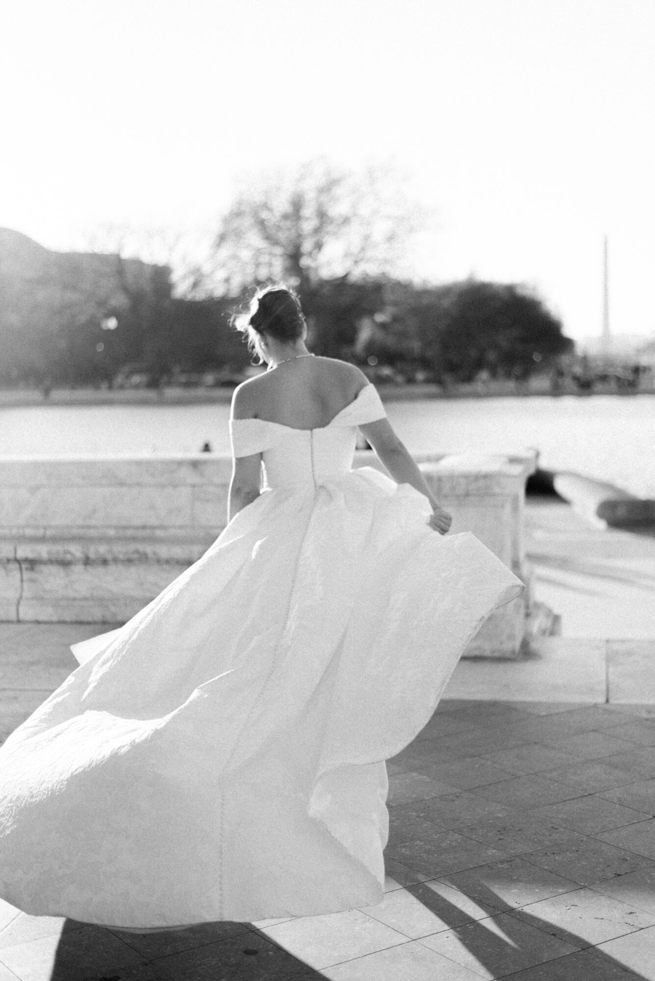 Woman in a flowing dress walking outdoors, back view, with trees and sky in background.