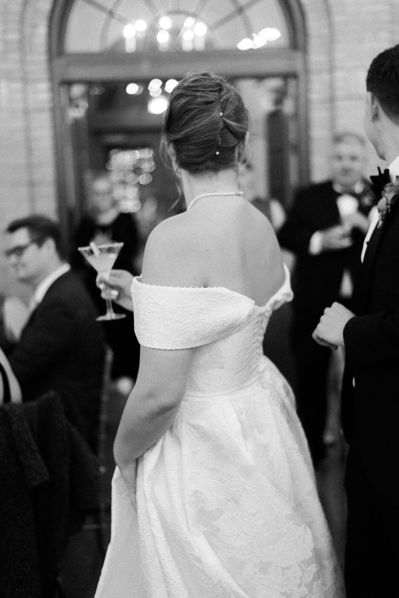 Woman in elegant dress with updo hairstyle at formal event, surrounded by people holding drinks, in a decorated indoor setting.