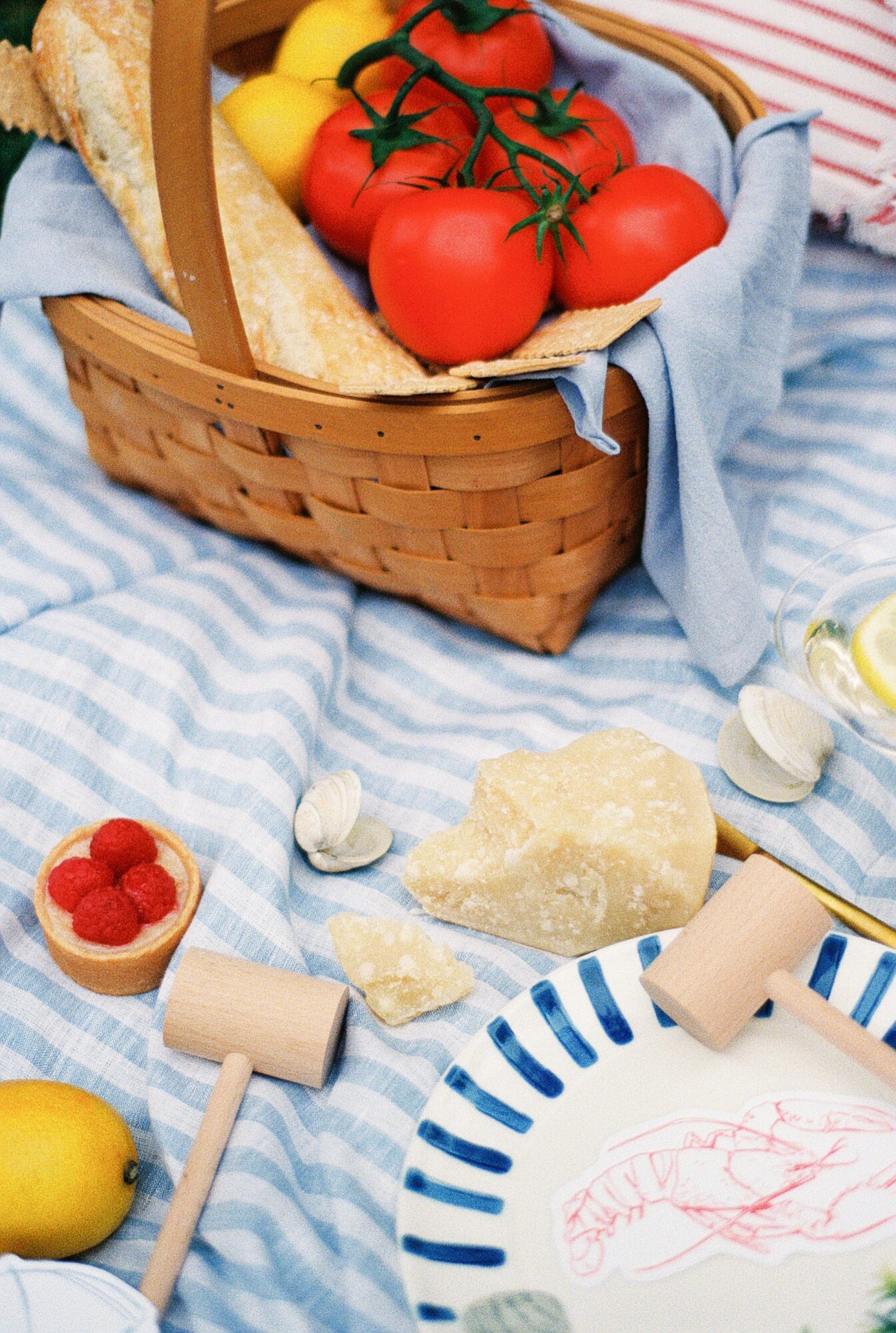 Basket with tomatoes and lemons, cheese, raspberries, and bread on a blue striped cloth, with a plate and utensils nearby.