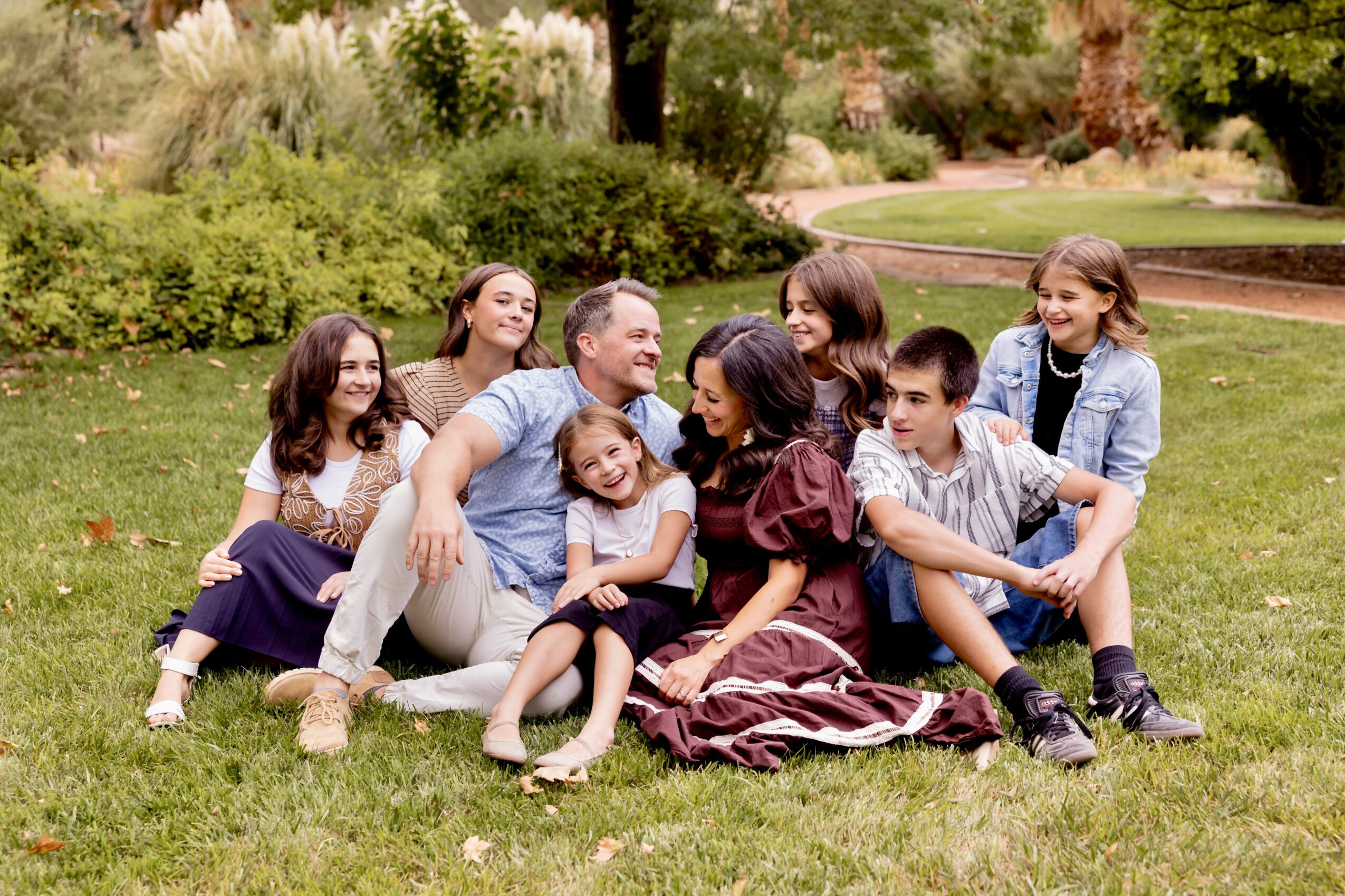 Group of seven people, including children and adults, sitting on grass in a park with trees and bushes in background.