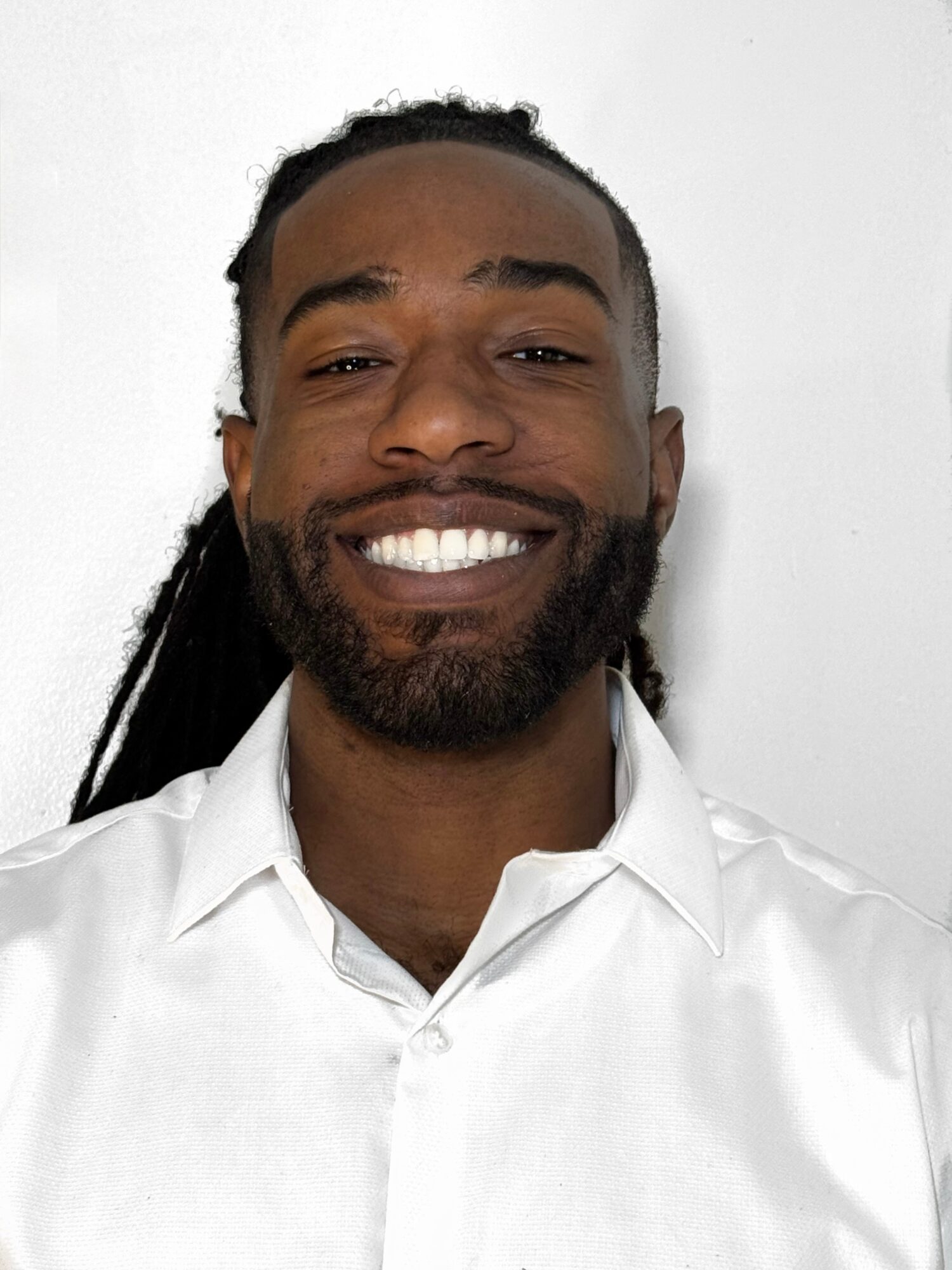 Smiling man with dreadlocks and beard wearing a white shirt against a plain background.