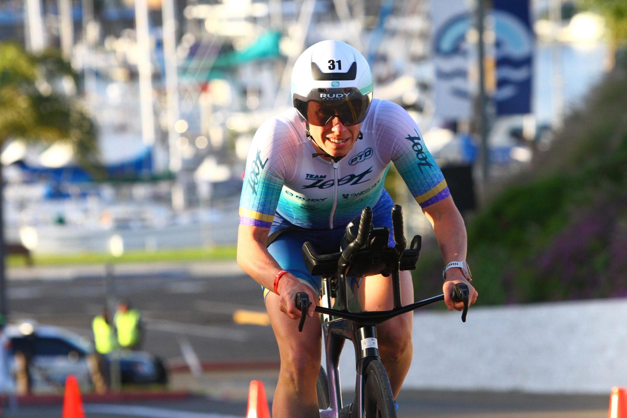 Cyclist wearing a helmet and sunglasses rides a bike outdoors with boats and water in the background.