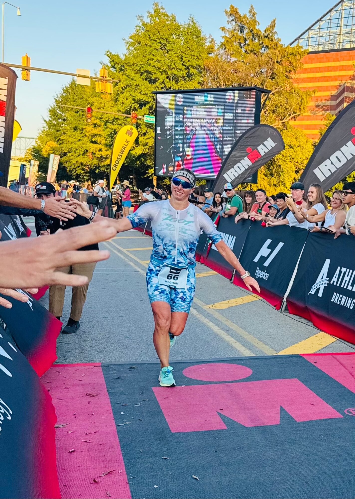 Female runner crossing finish line with outstretched arms, smiling, wearing sunglasses and blue athletic outfit, crowd cheering nearby.