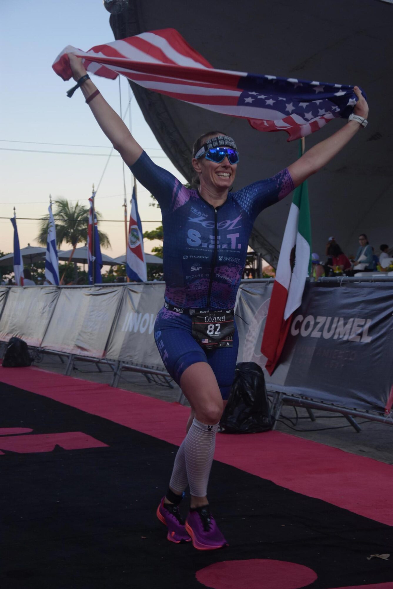 Woman celebrating with an American flag, wearing sunglasses, a blue athletic outfit, and pink running shoes, on a red carpet.