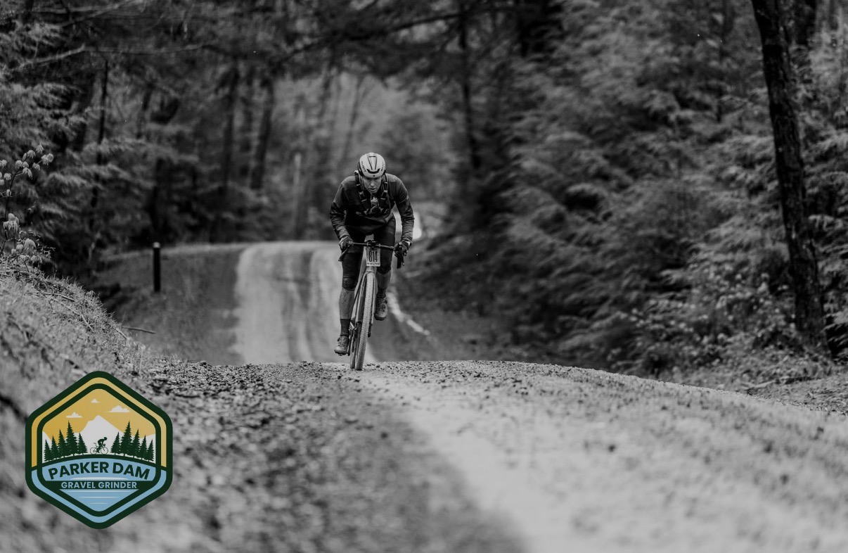 Person riding a mountain bike on a dirt trail through a forest, black and white image, Parker Dam logo in bottom left corner.