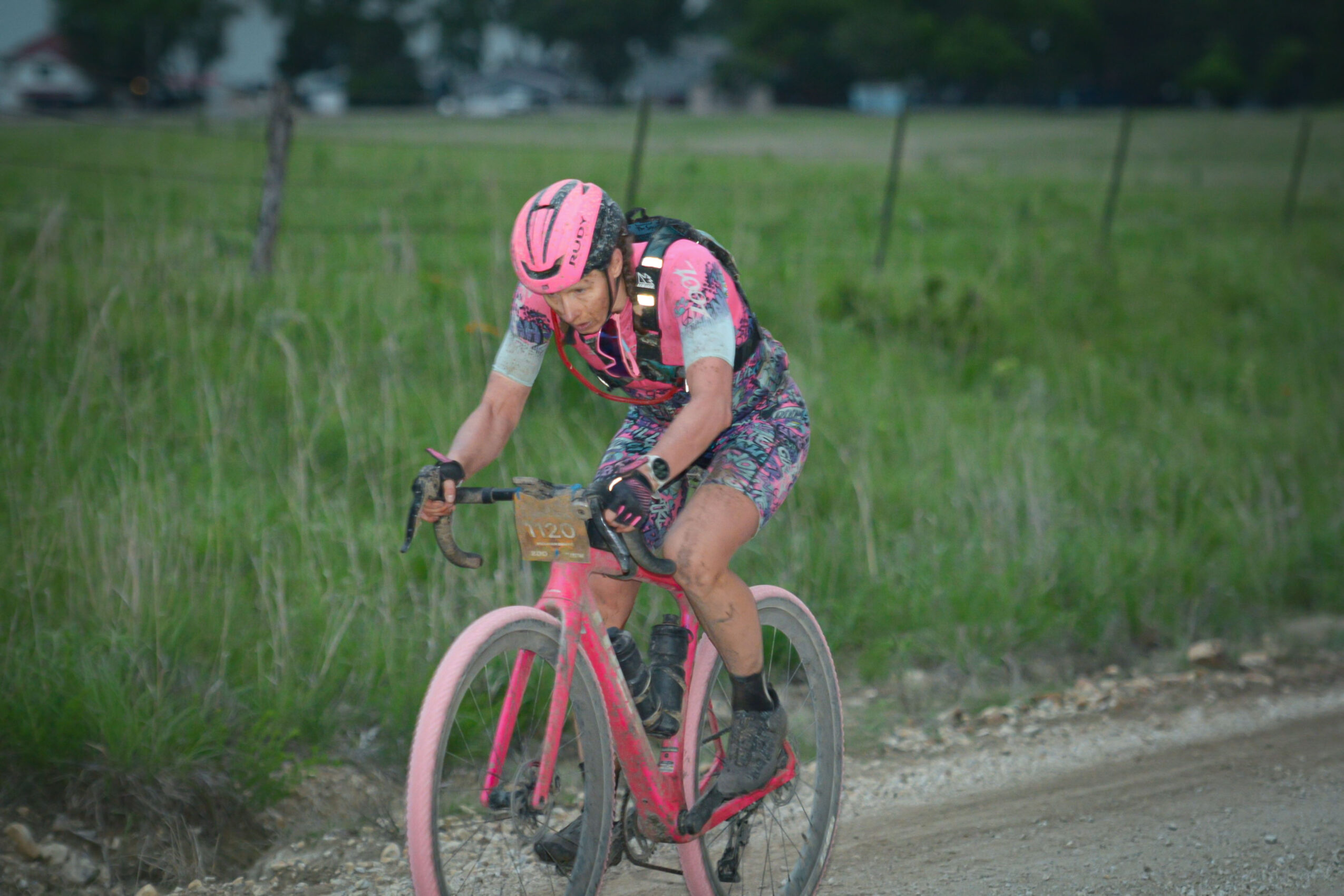 Person riding a pink bicycle on a dirt path in a grassy field, wearing a pink helmet and colorful clothing.