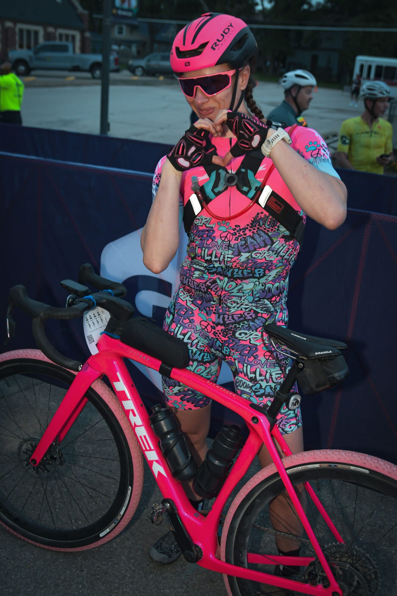 Person in cycling gear, pink helmet, sunglasses, standing with pink bicycle, outdoors, other cyclists in background.