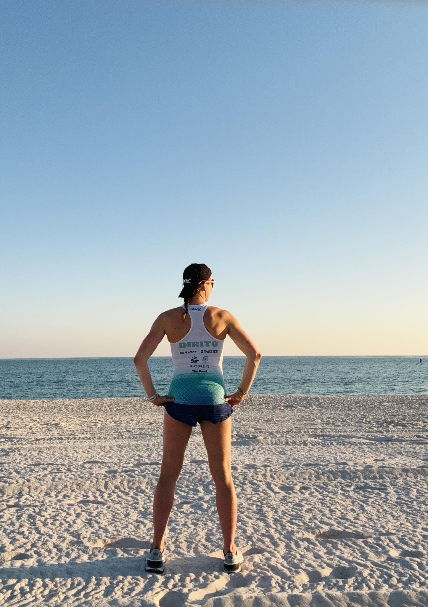 Person standing on sandy beach facing the ocean with hands on hips, wearing a tank top and shorts, under a clear blue sky.