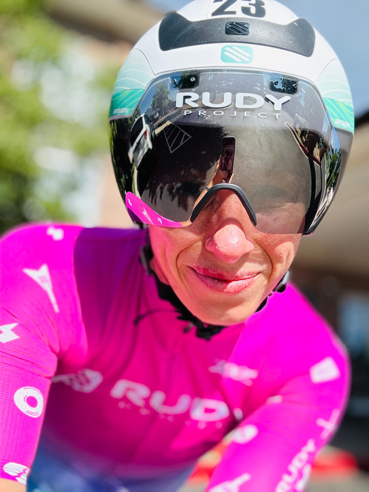 Close-up of a cyclist wearing a white helmet with black and teal accents, pink cycling jersey, and reflective sunglasses.
