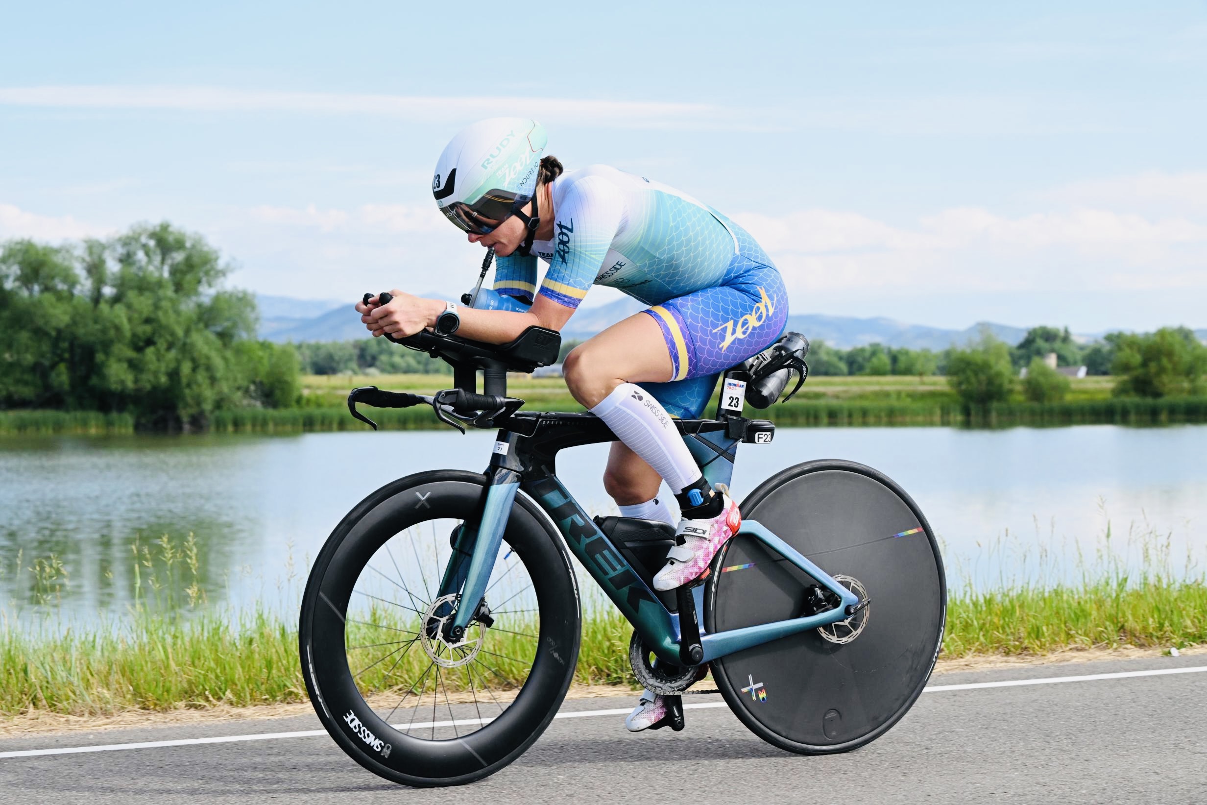 Cyclist wearing a helmet and sportswear rides a bike along a lakeside path with trees and water in the background.