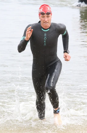 Person running out of water wearing a wetsuit, red swim cap, and goggles, with water splashing around.