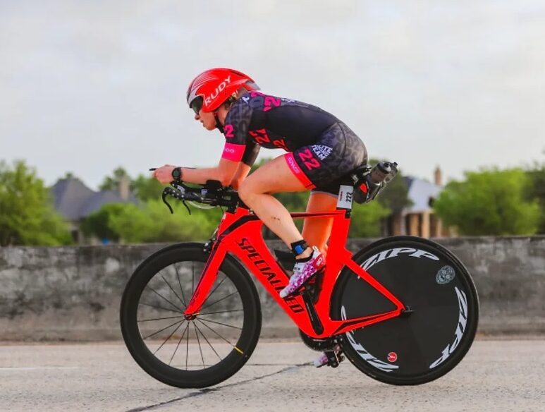 Cyclist in black and pink gear riding a red racing bike on a road with trees and sky in background