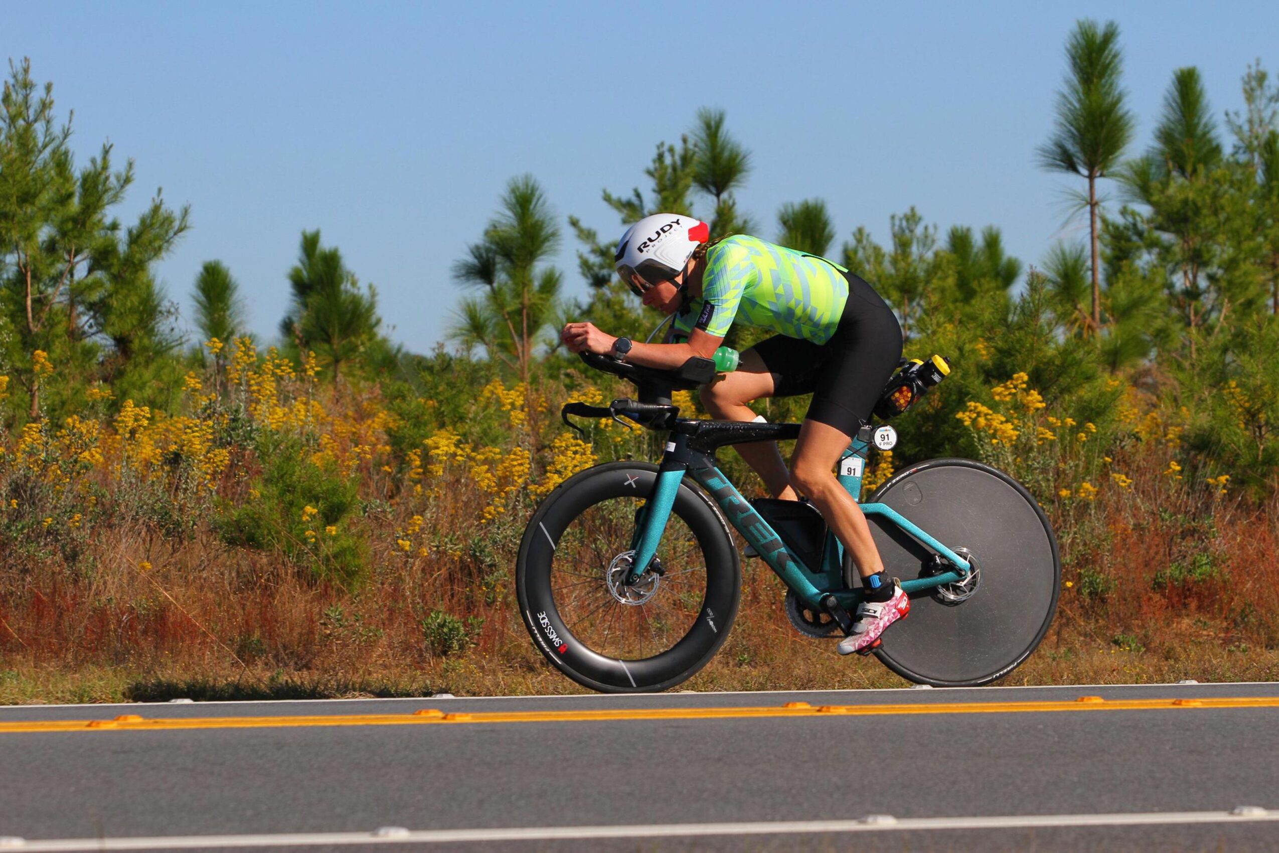 Person riding a bicycle on a road with trees and yellow flowers in the background, wearing a helmet and bright clothing.