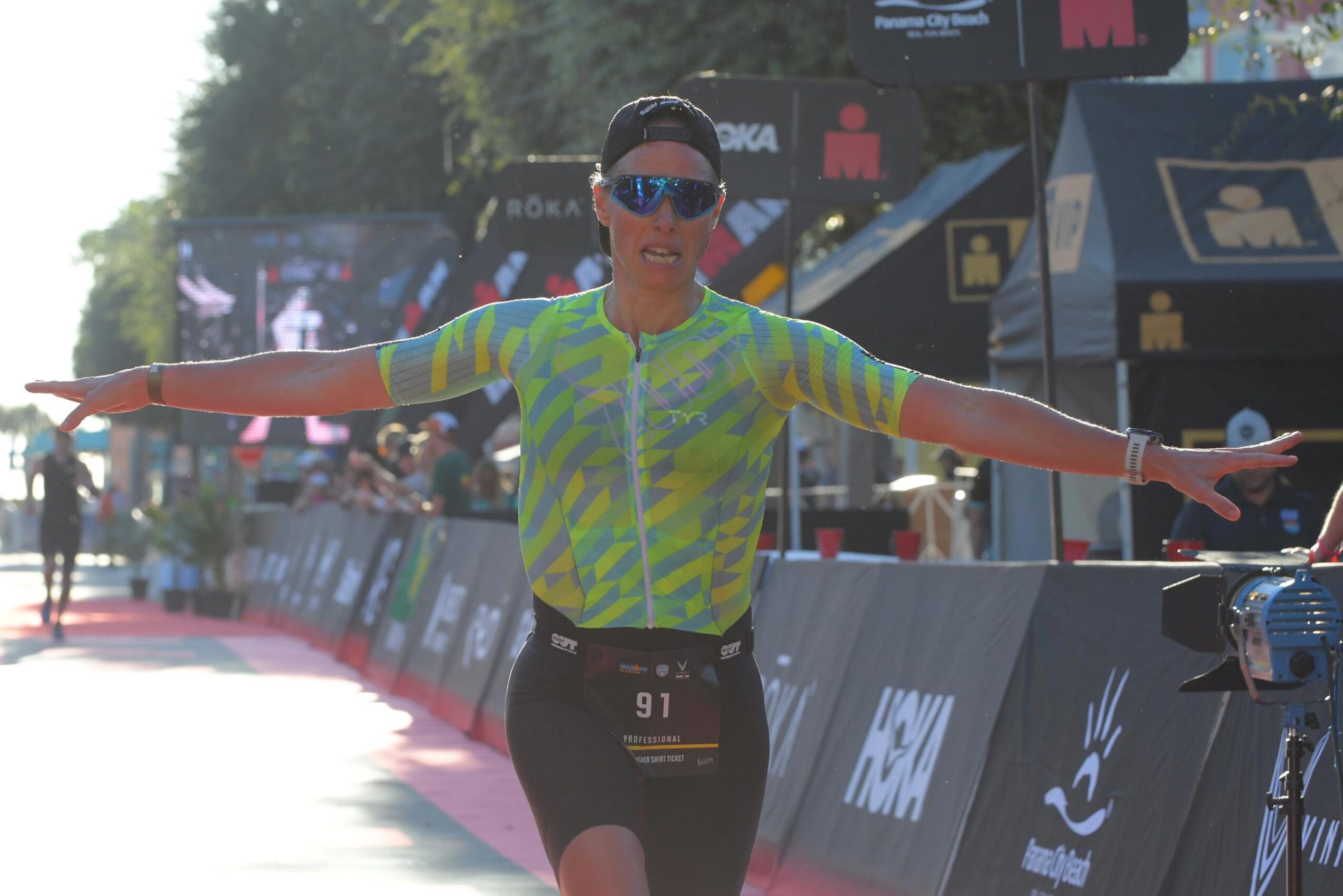 Female athlete with arms outstretched, wearing sunglasses and a colorful shirt, standing on a race course with banners and spectators.