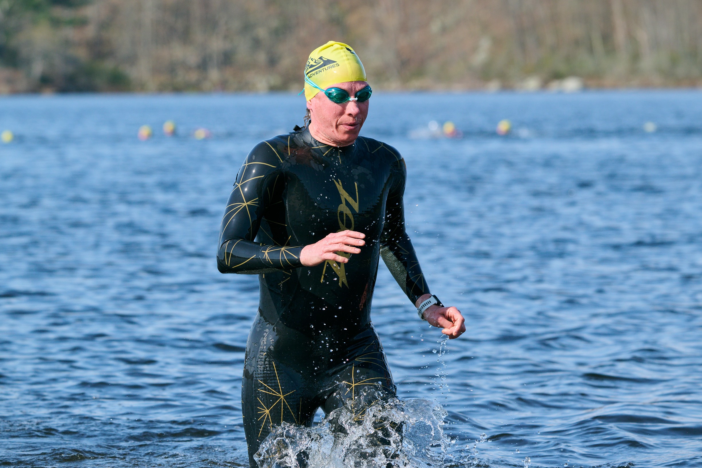 Person in a wetsuit running in water, wearing goggles and a yellow swim cap, with a lake and trees in background.